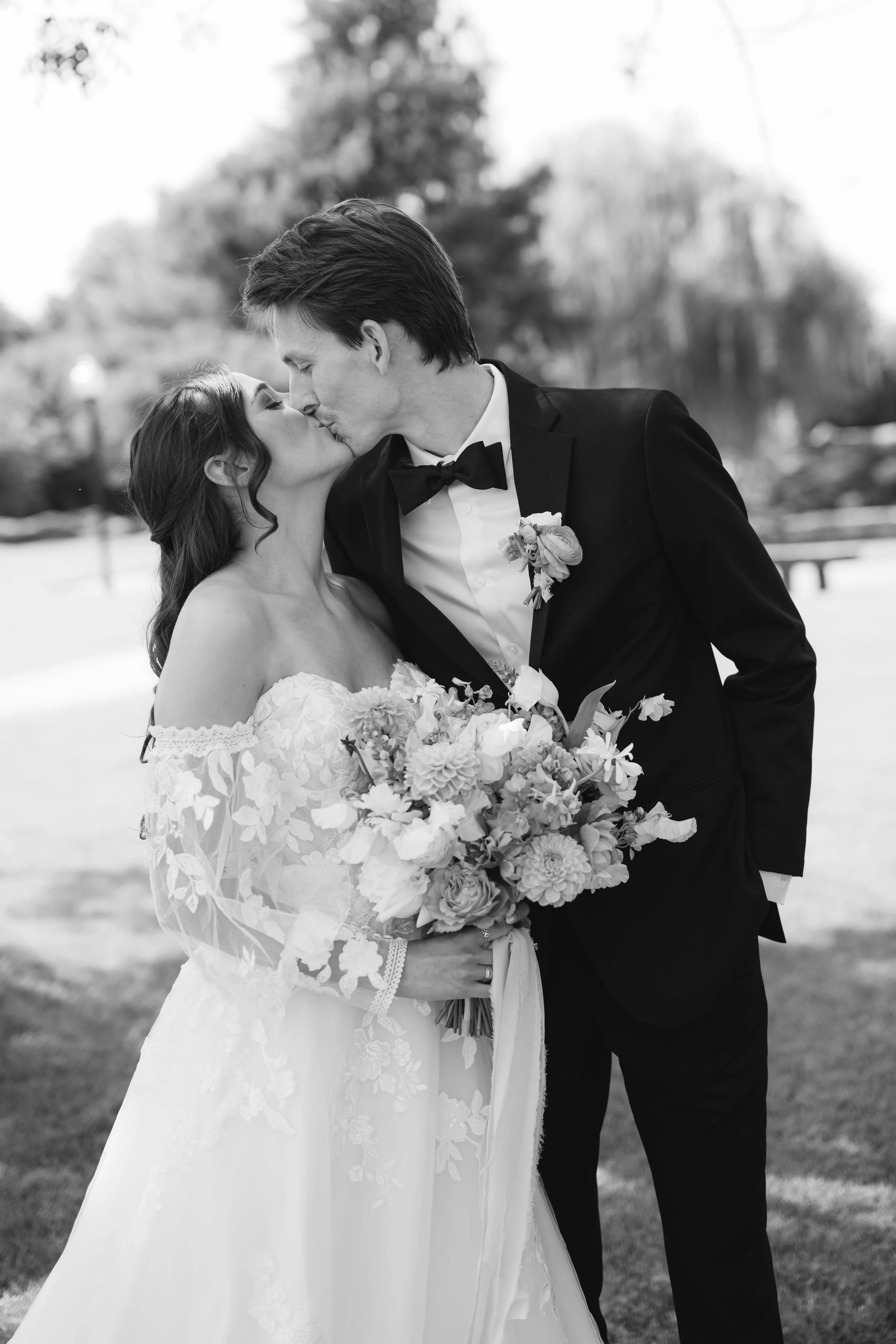 Black and white photo of a bride and groom kissing outdoors, the bride holding a bouquet of flowers.