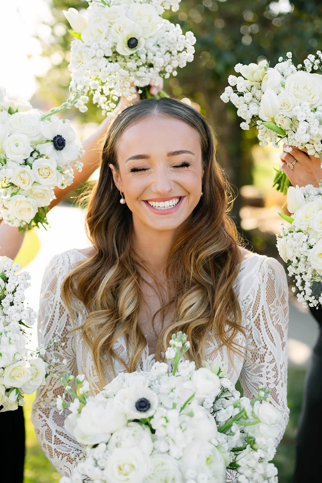 A smiling woman with long wavy hair, wearing a lace dress and pearl earrings, is surrounded by white floral bouquets with an outdoor background. She appears to be at a wedding or special event.