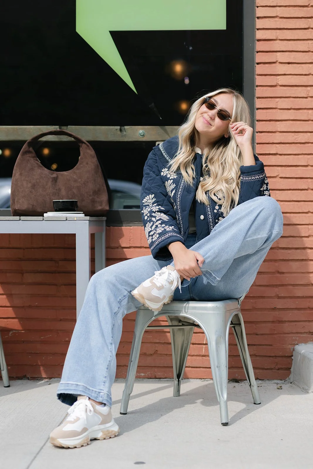 Young woman with long blonde hair wearing sunglasses, a navy blue embroidered jacket, and light blue jeans, sitting casually on a metal chair outside a building with brick wall, crossing her leg and smiling.