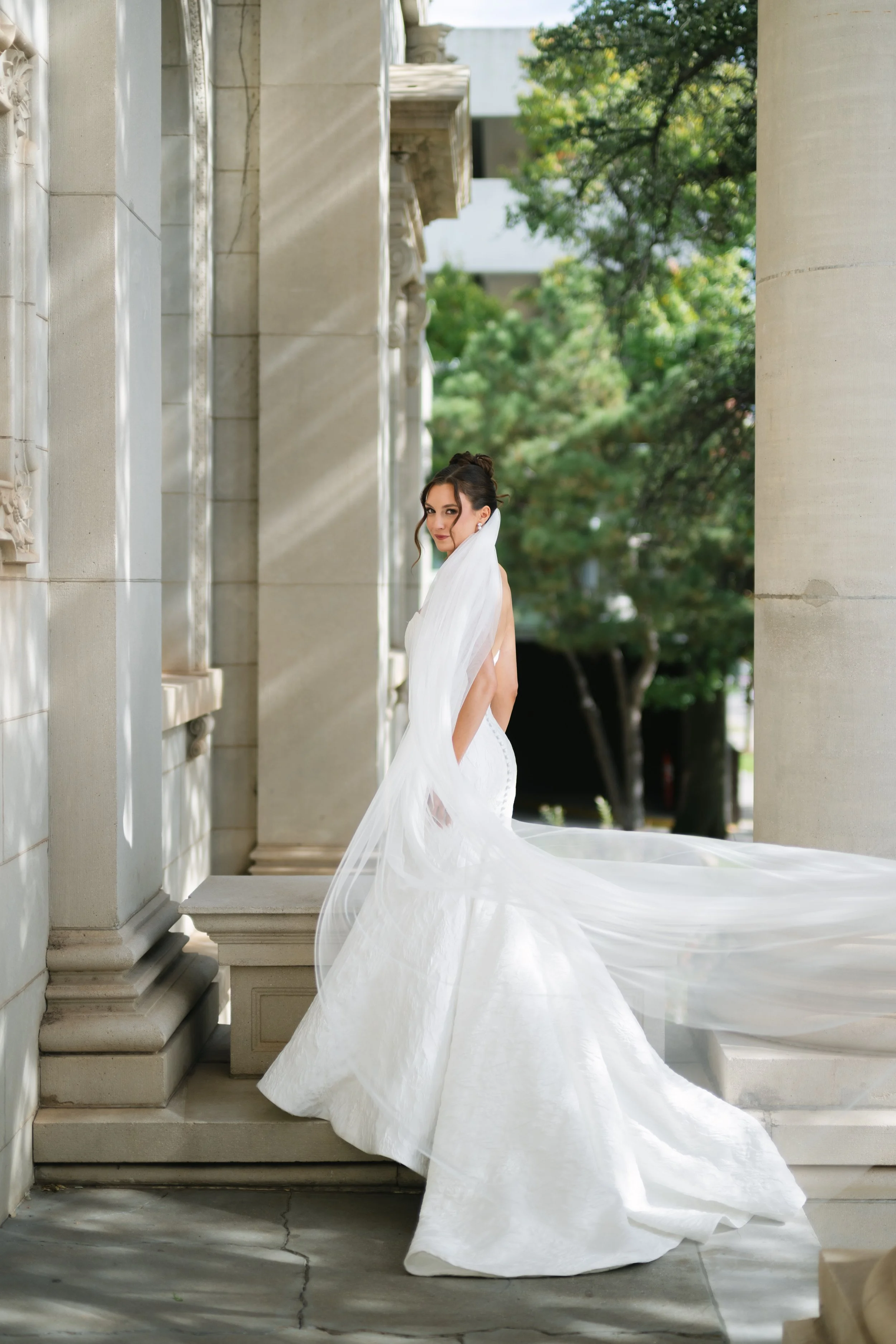 A woman in a white wedding dress with a long train and veil standing on the steps of a stone building with columns, outdoors with trees in the background.