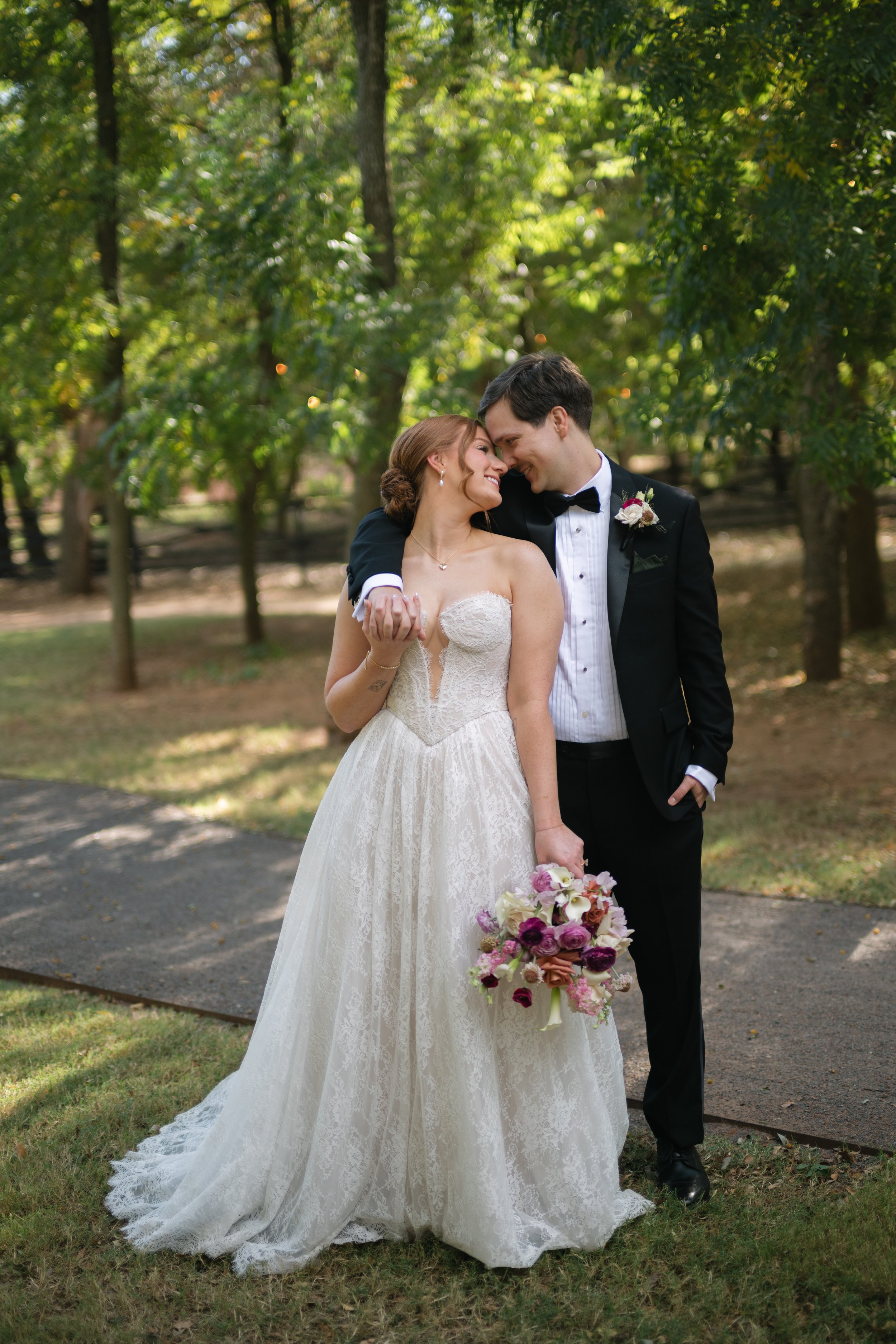 A newlywed couple is standing closely together outdoors among trees, smiling and touching foreheads. The bride is wearing a strapless lace wedding gown and holding a bouquet of pink, purple, and white flowers. The groom is dressed in a black tuxedo with a bow tie and a boutonniere. The couple appears happy and romantic.