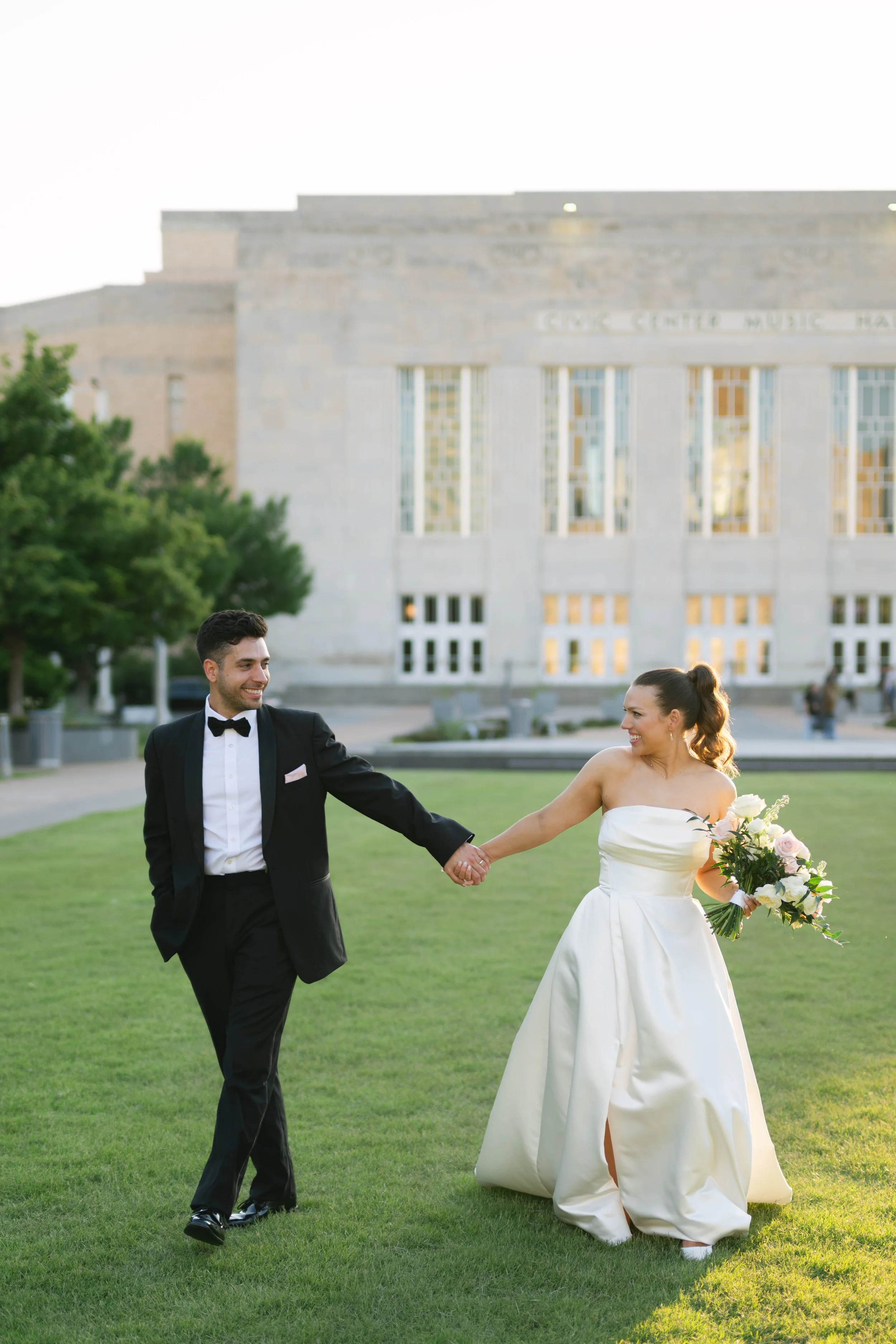 A bride and groom holding hands and walking outdoors on a grassy area with a historic building in the background.