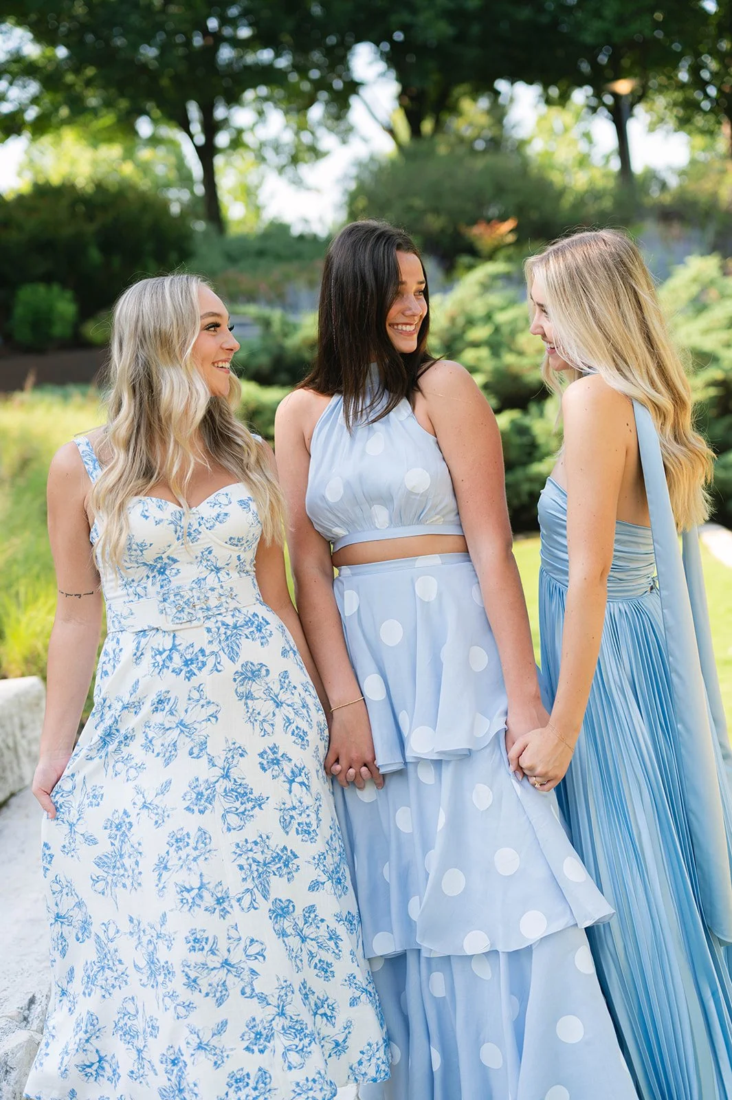Three women wearing pastel blue dresses are standing outdoors, holding hands and smiling at each other, with a green garden and trees in the background.