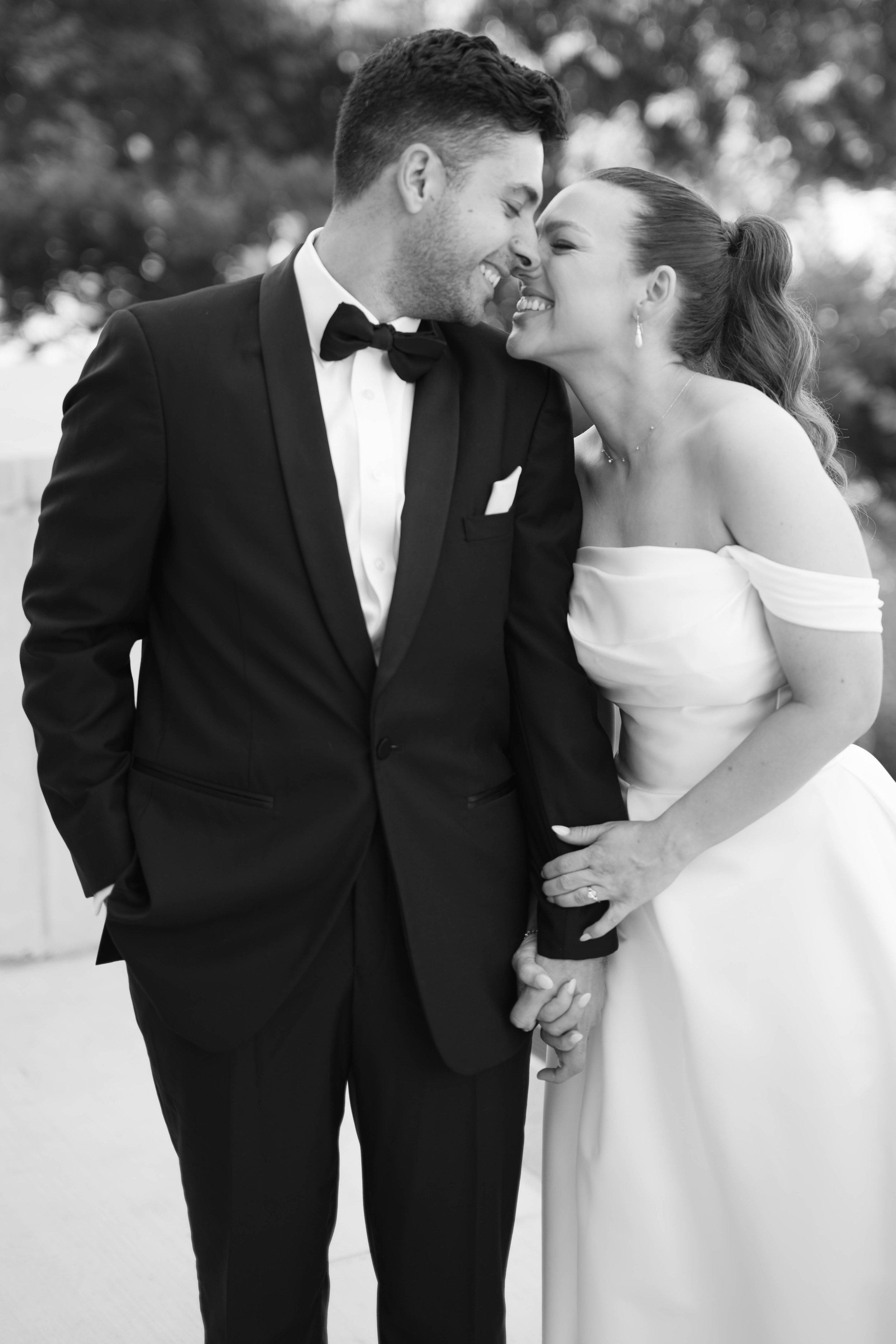 Black and white photo of a bride and groom smiling and touching foreheads outside, holding hands, dressed in wedding attire.
