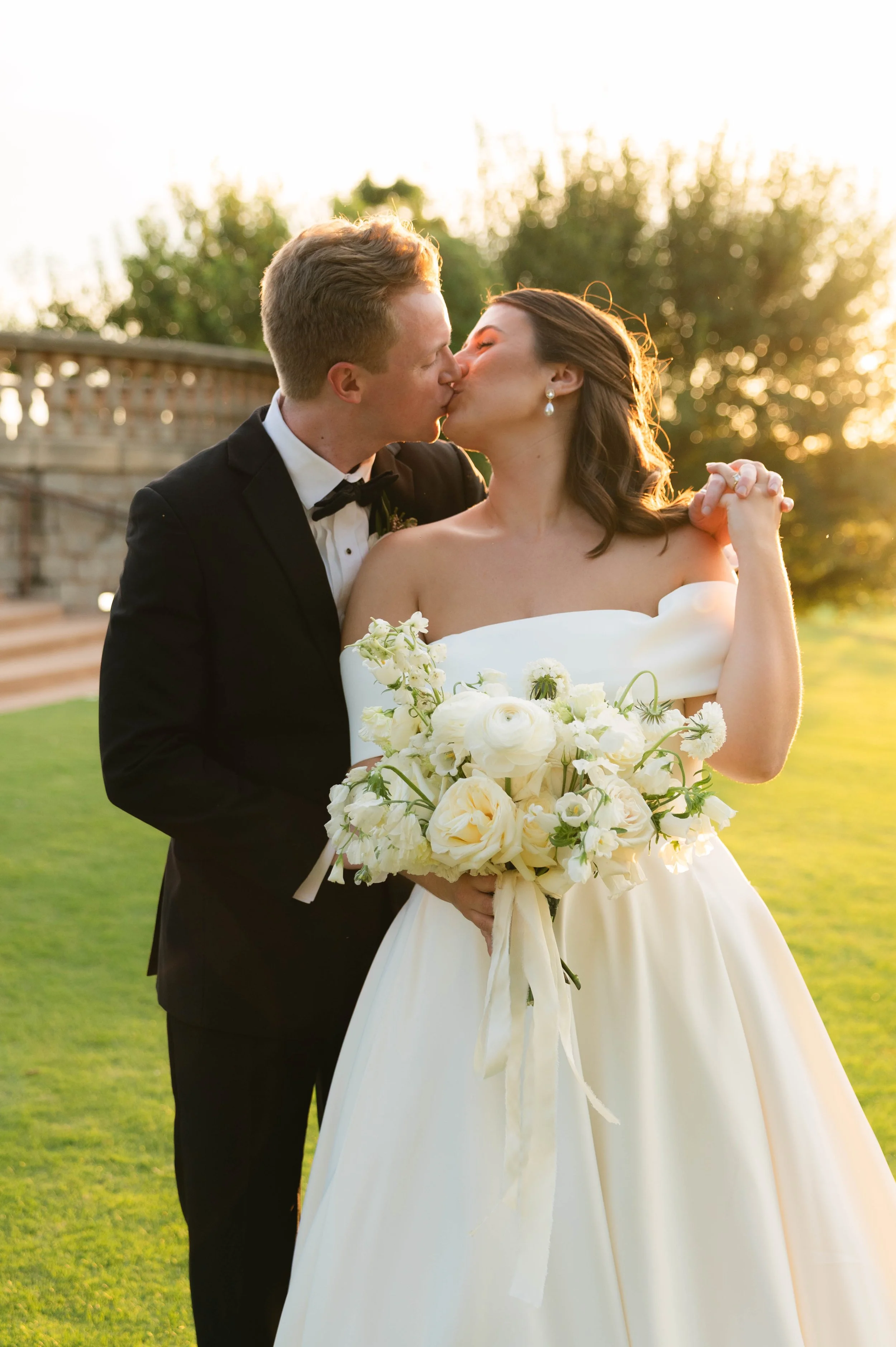 A bride and groom kiss outdoors at sunset, with the bride holding a bouquet of white flowers, in a park or garden setting.