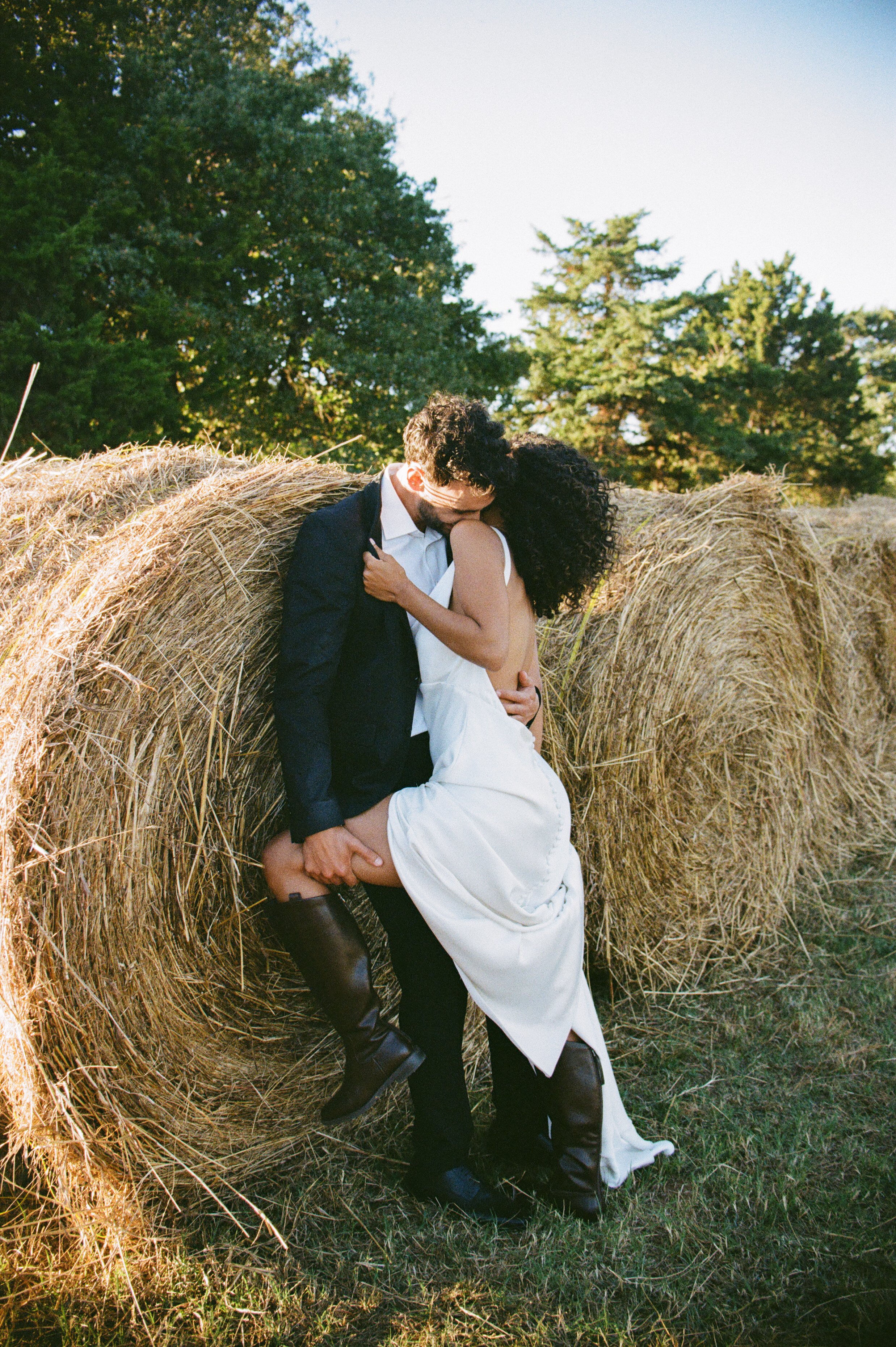 A man and woman embracing in a field with hay bales and trees in the background