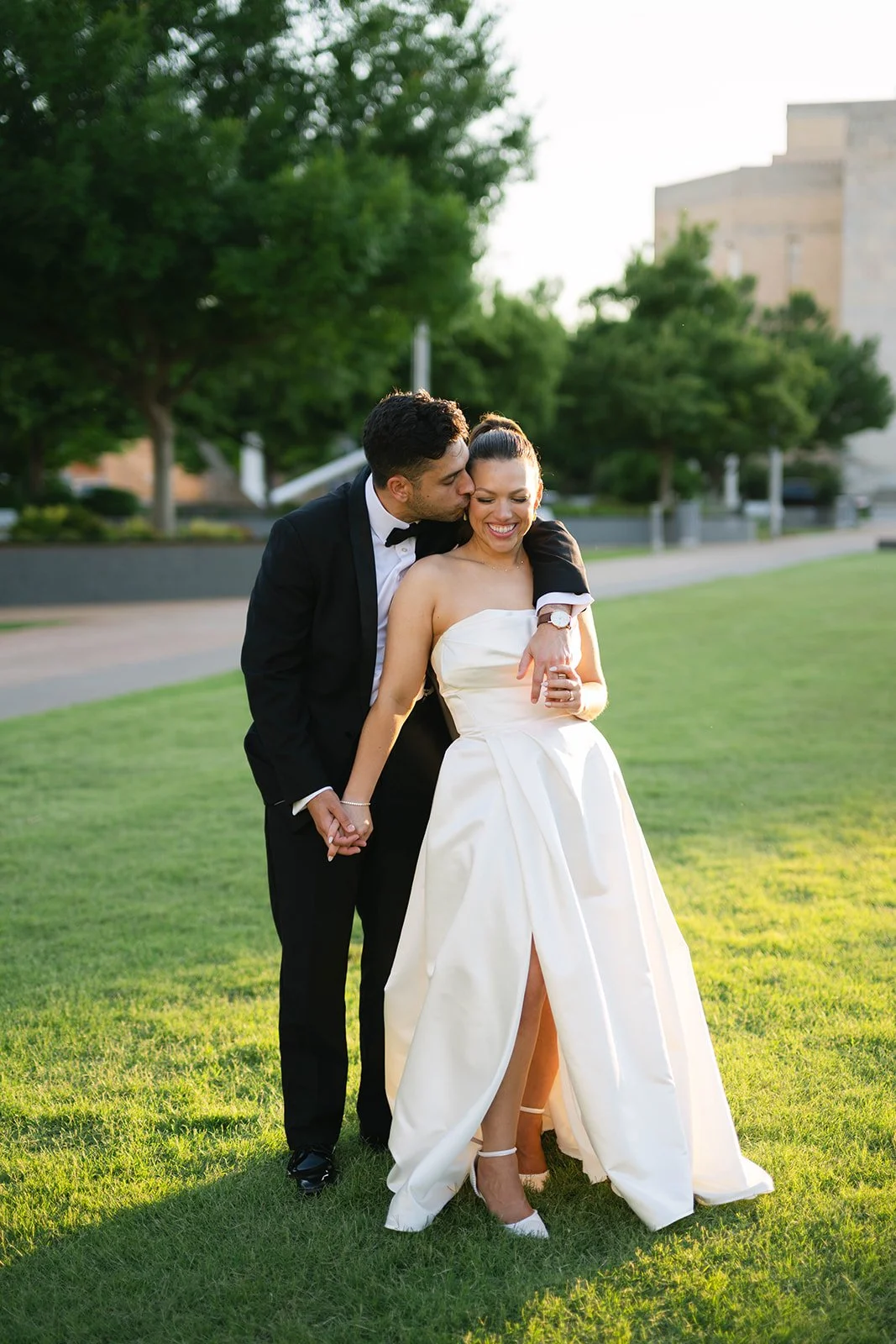 A newlywed couple holding hands and sharing a kiss in a park, with the bride in a strapless white gown and the groom in a black tuxedo.