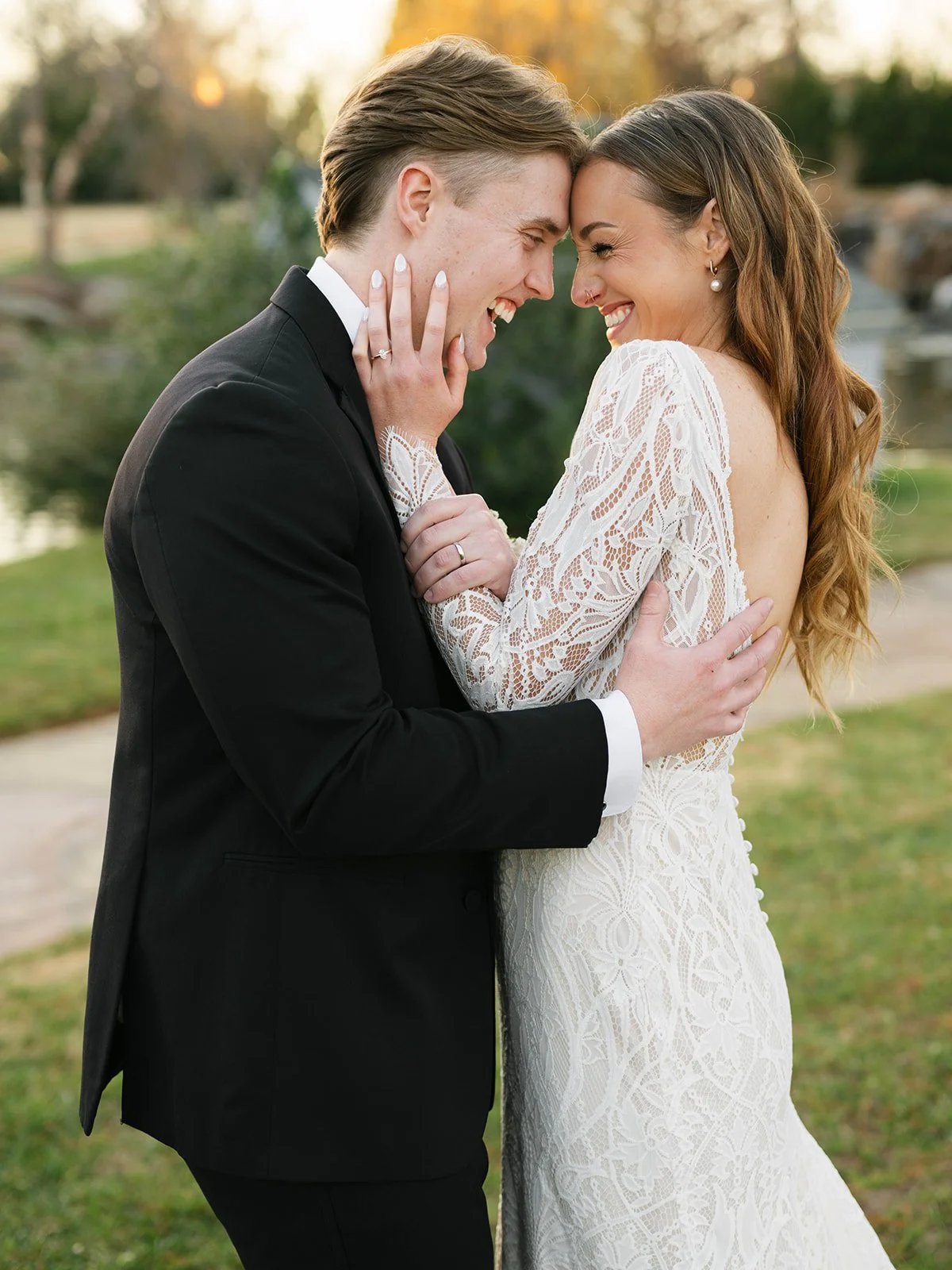 A couple in wedding attire sharing a joyful moment, touching foreheads and smiling, outdoors during sunset.