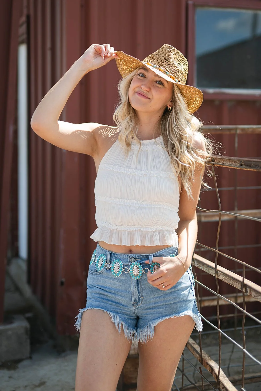 A young woman in a white sleeveless crop top, denim shorts with a floral belt, and a wide-brimmed straw hat, standing outdoors near a metal fence and red wooden building.