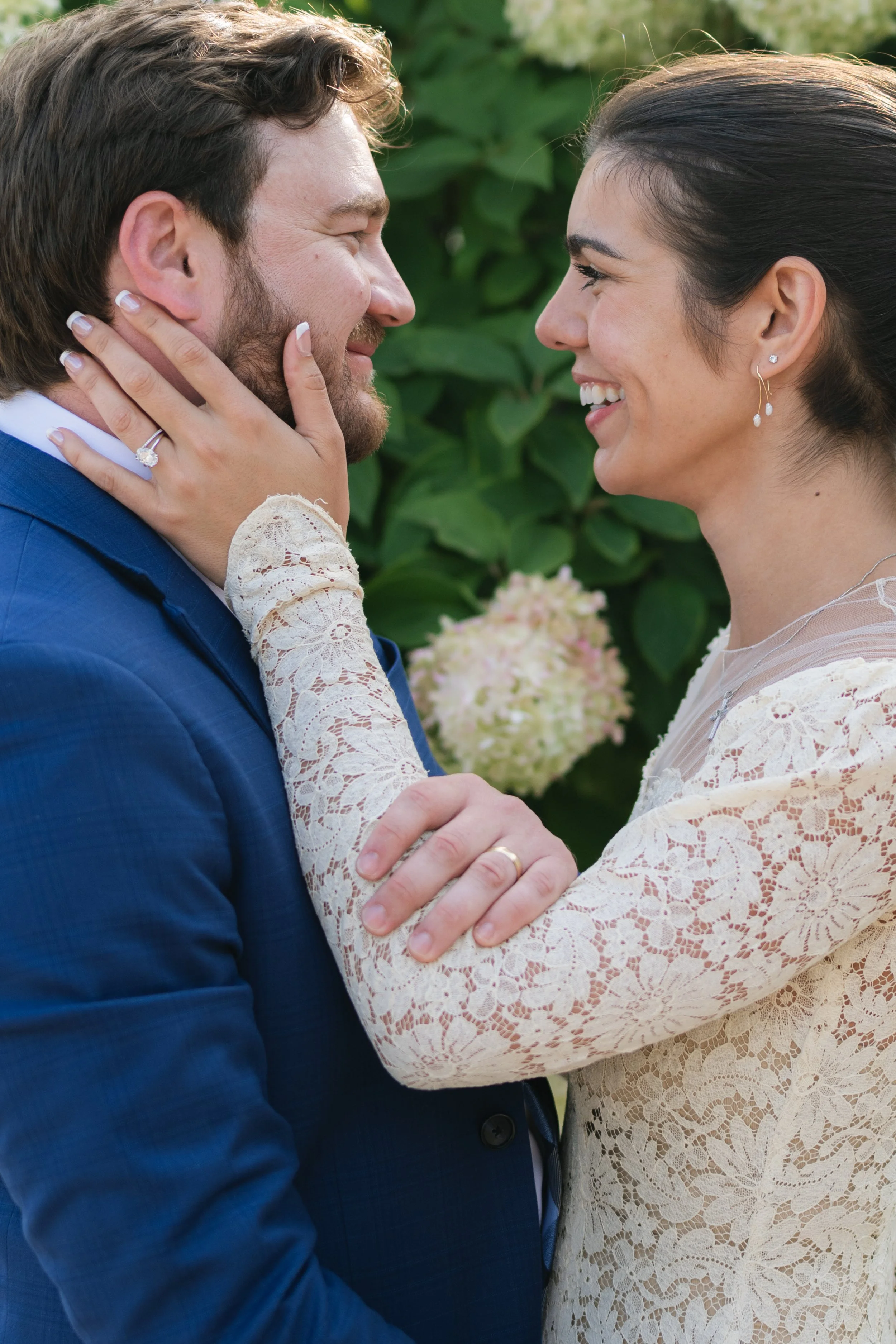 A couple on their wedding day, looking into each other's eyes with smiles, outdoors with green foliage in the background.