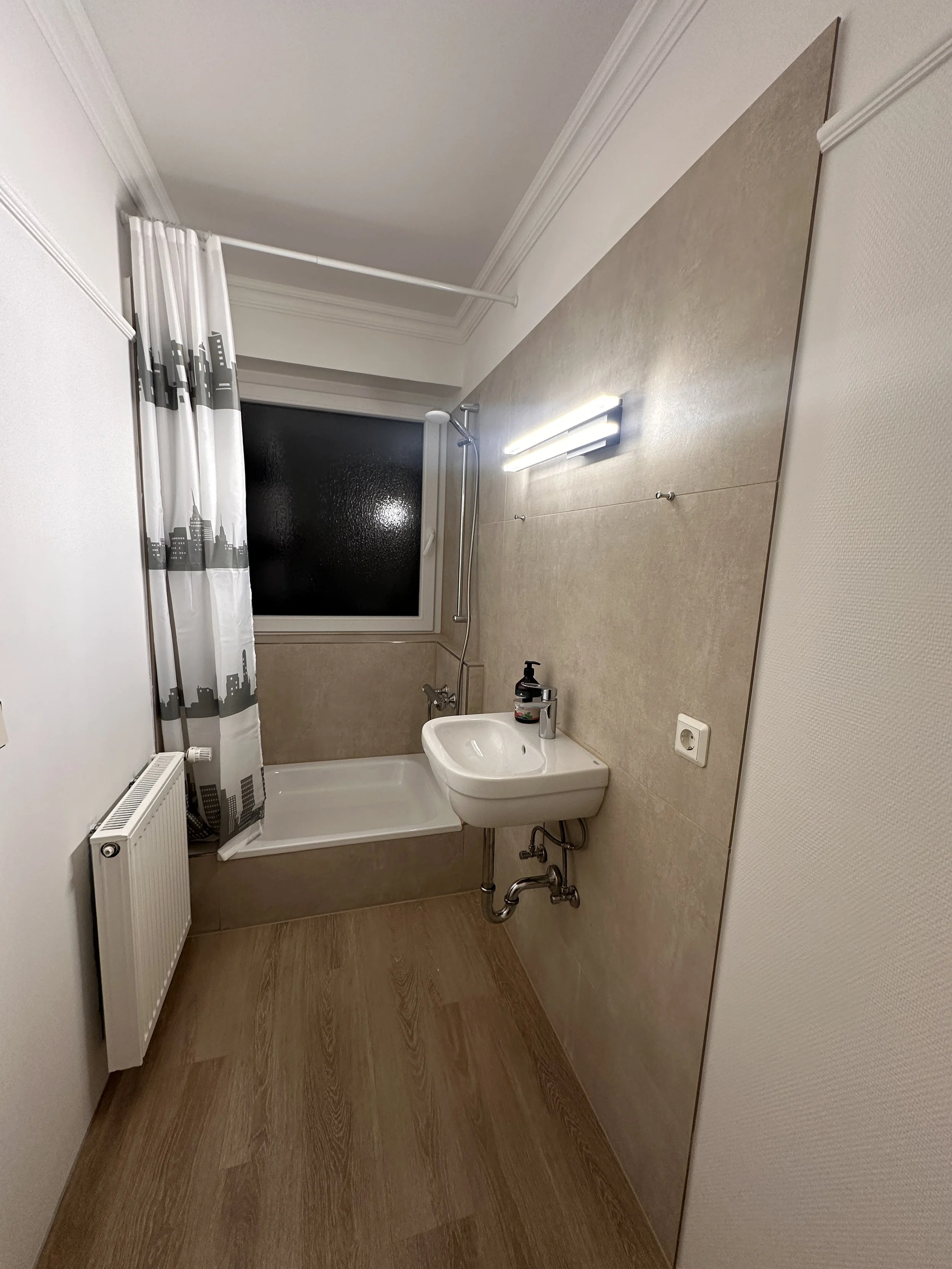 Kleines Badezimmer mit Badewanne, Waschbecken, dunklem Fenster, weißen Wänden, Holzboden, Heizkörper, und gestrichener Wand mit Licht.