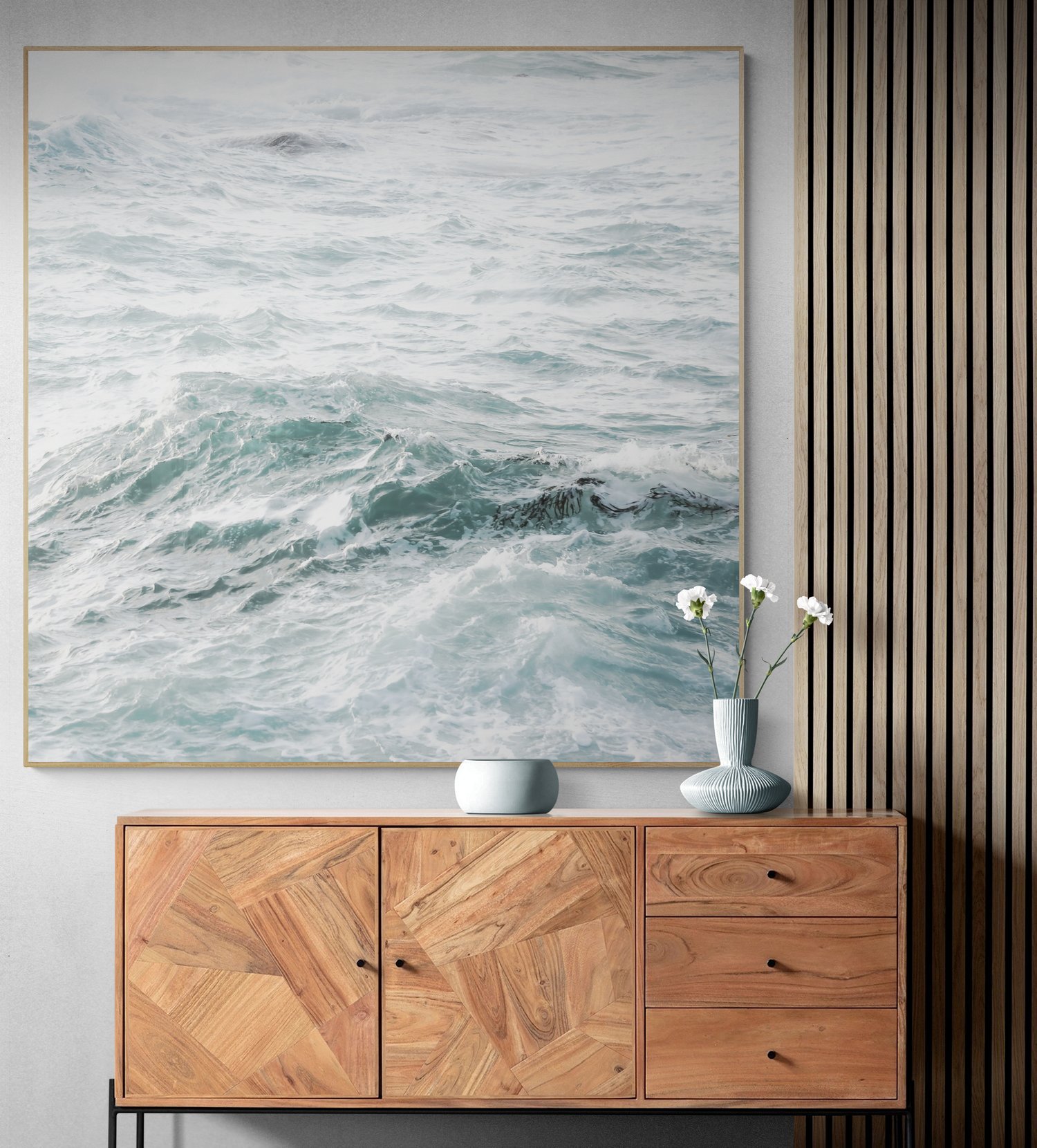 Interior design featuring a large ocean wave painting above a wooden sideboard. The sideboard has a geometric pattern and supports two vases, one with white flowers. Vertical wood paneling is visible on the right wall.