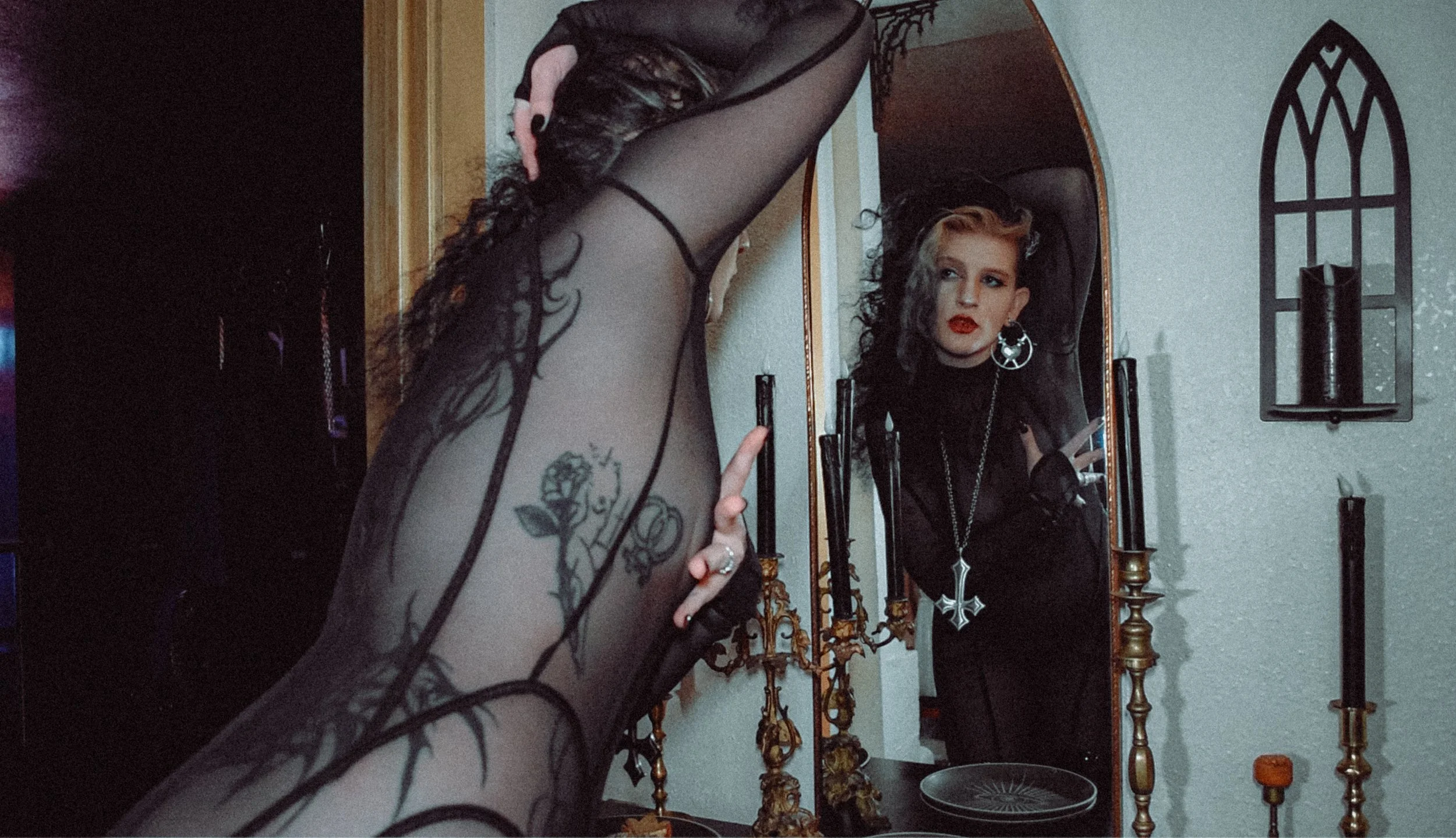 A dominant woman gazes into a mirror clad in a black catsuit, their tattoos visible through the mesh