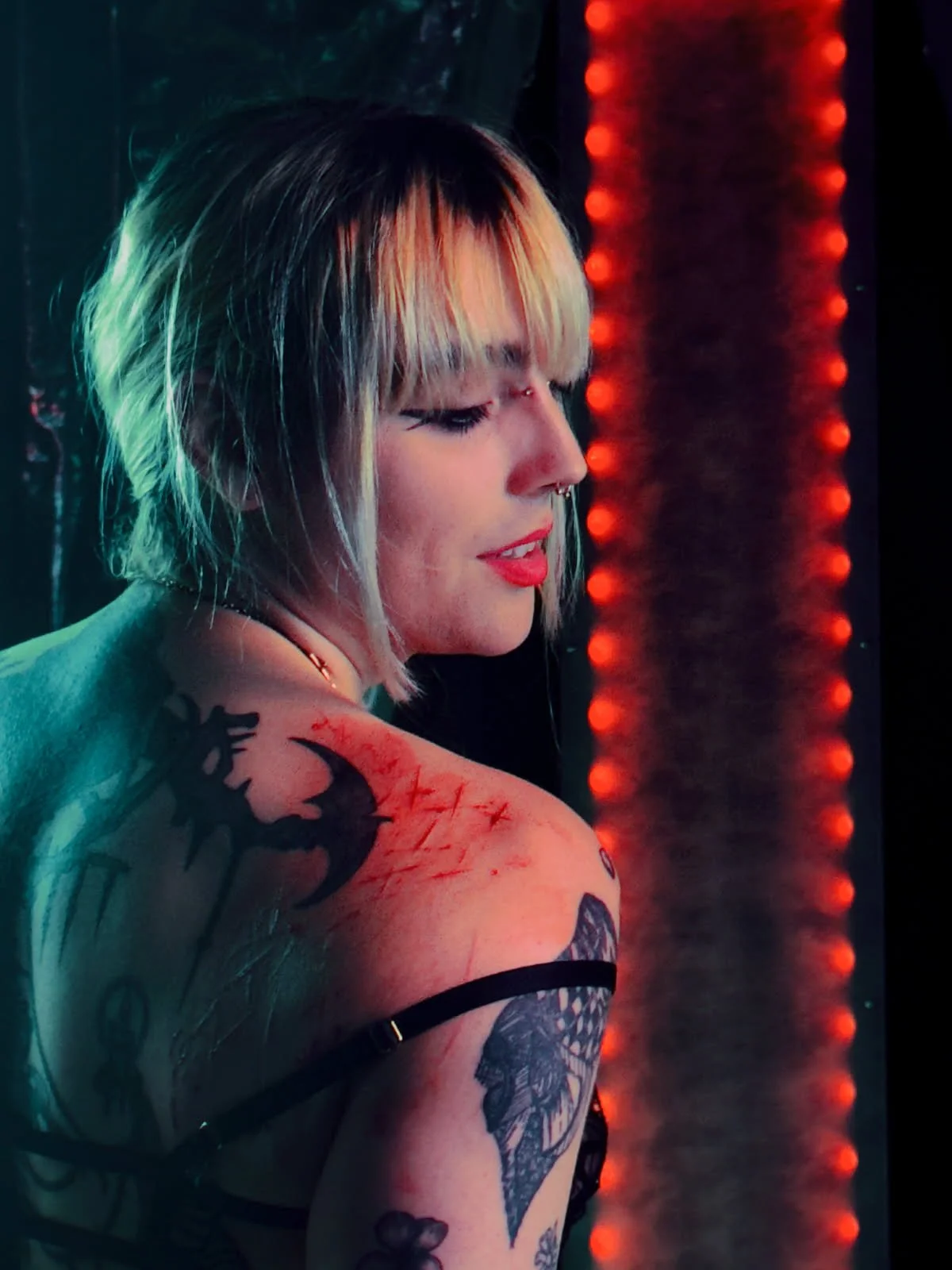 A young woman with short, blond hair and multiple tattoos looks to the side. She has a septum piercing and is wearing visible black lingerie straps. The background features a dark setting with red-orange string lights.
