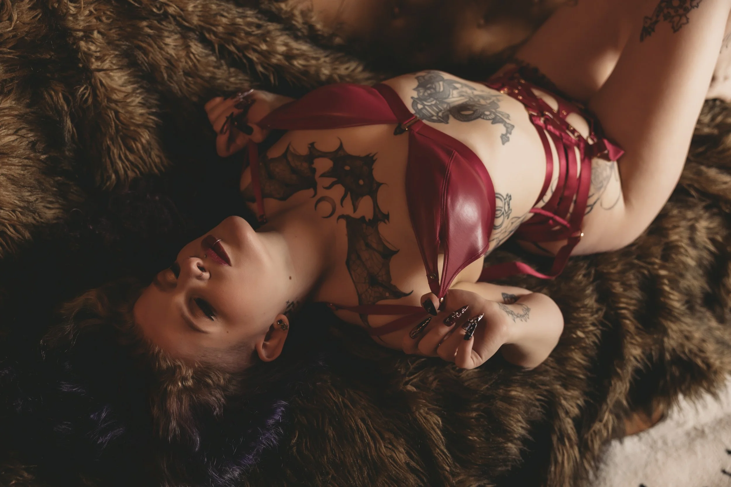 A woman lying on a fur rug, wearing red lingerie with multiple tattoos visible on her body.