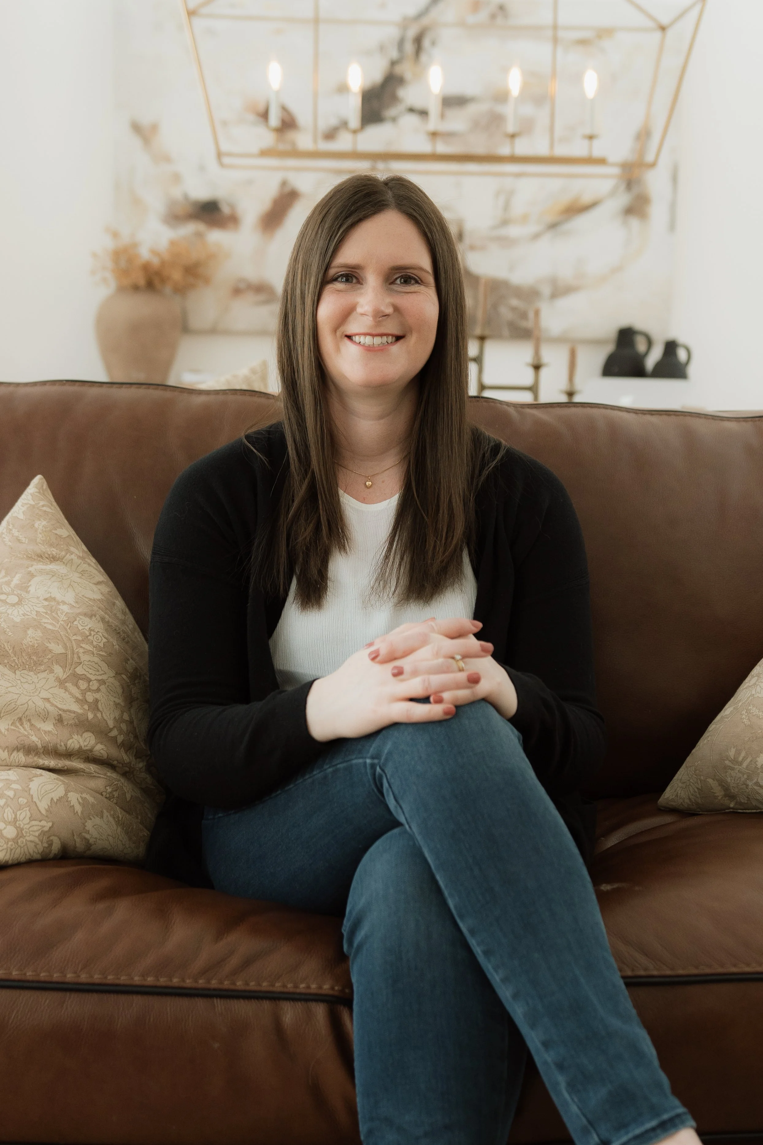 A photo of Rachelle Stephens, owner of Rachelle Stephens Counselling & Consulting - a woman with long brown hair smiling and sitting cross-legged on a brown leather couch.