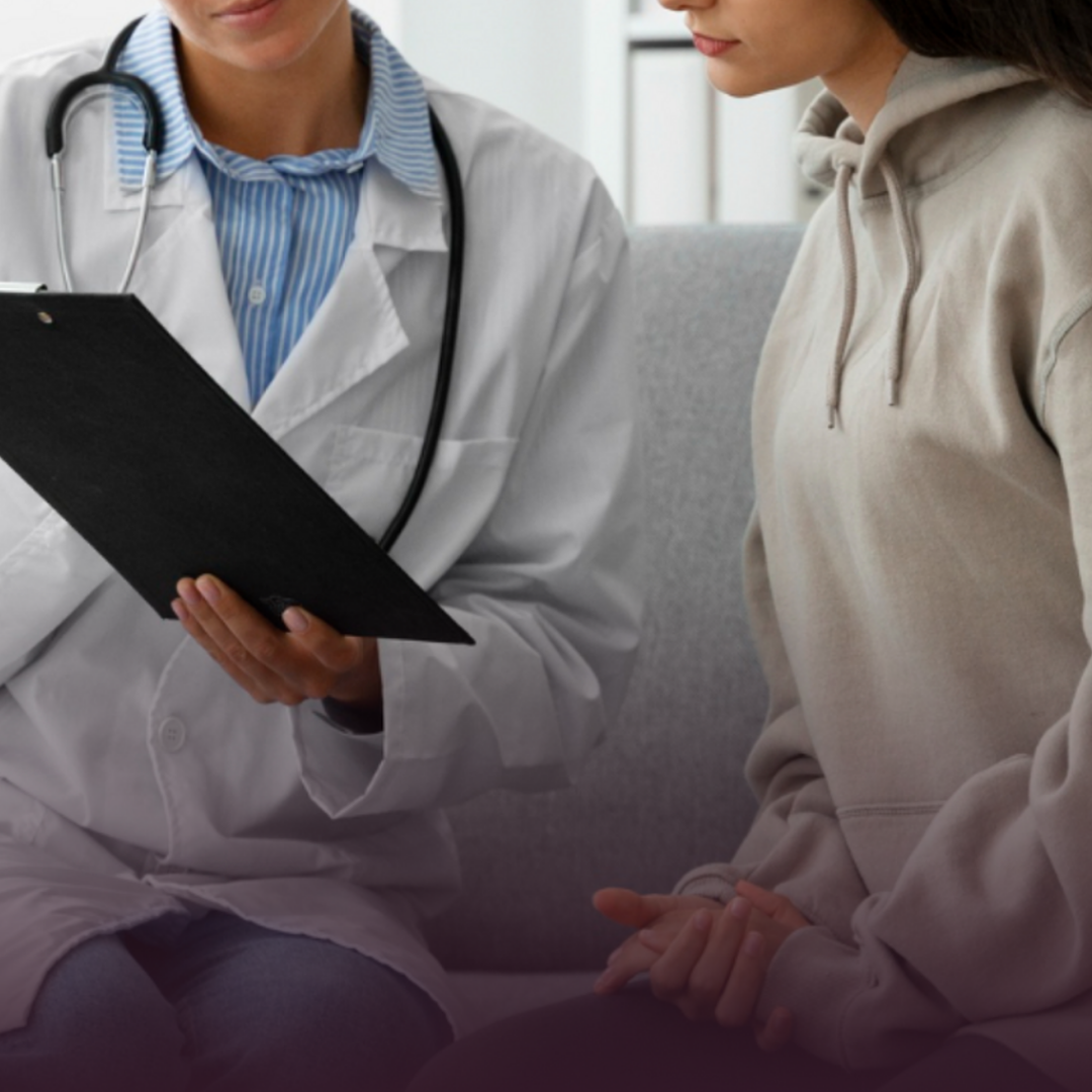 A healthcare professional in a white coat and stethoscope consults with a female patient in a beige hoodie in a medical setting.