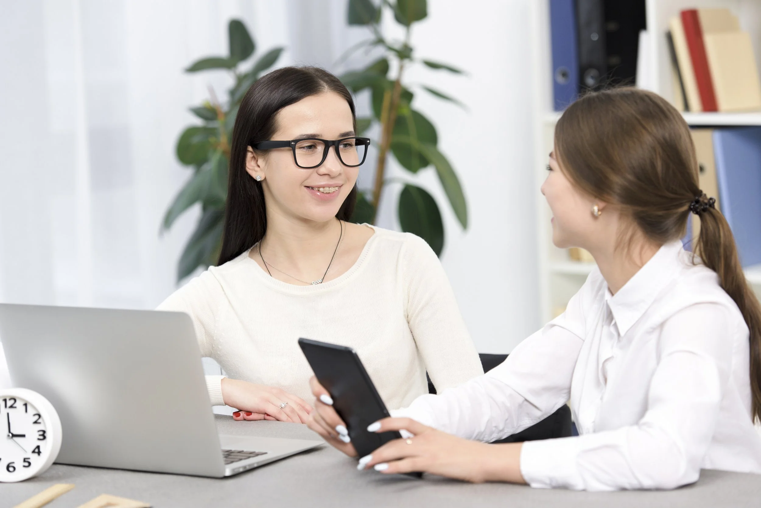 Two women sitting at a desk, one showing something on her phone to the other, with plants and shelves in the background.
