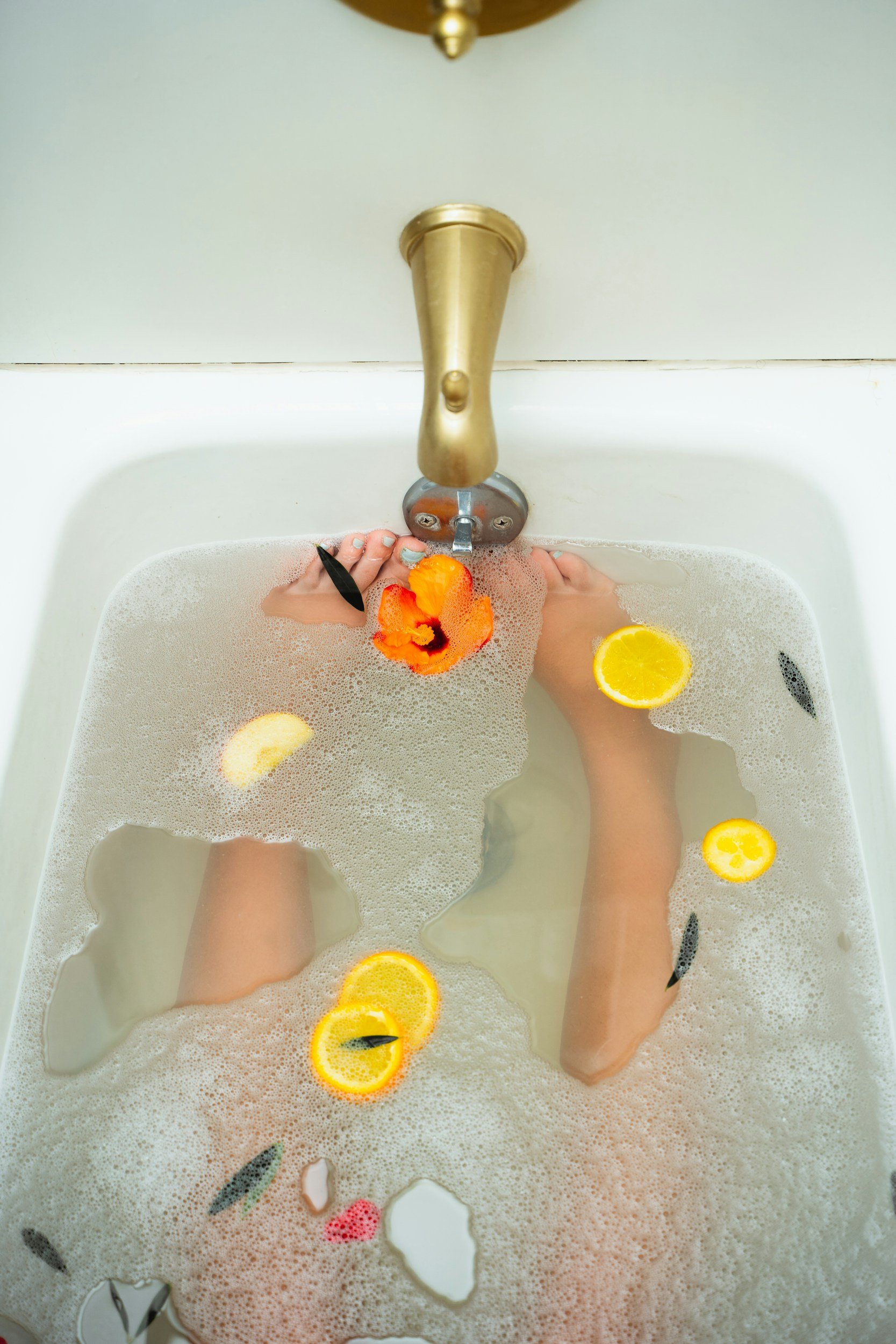 Feet in a bubble bath with lemon slices, flower, and small fish, viewed from above.