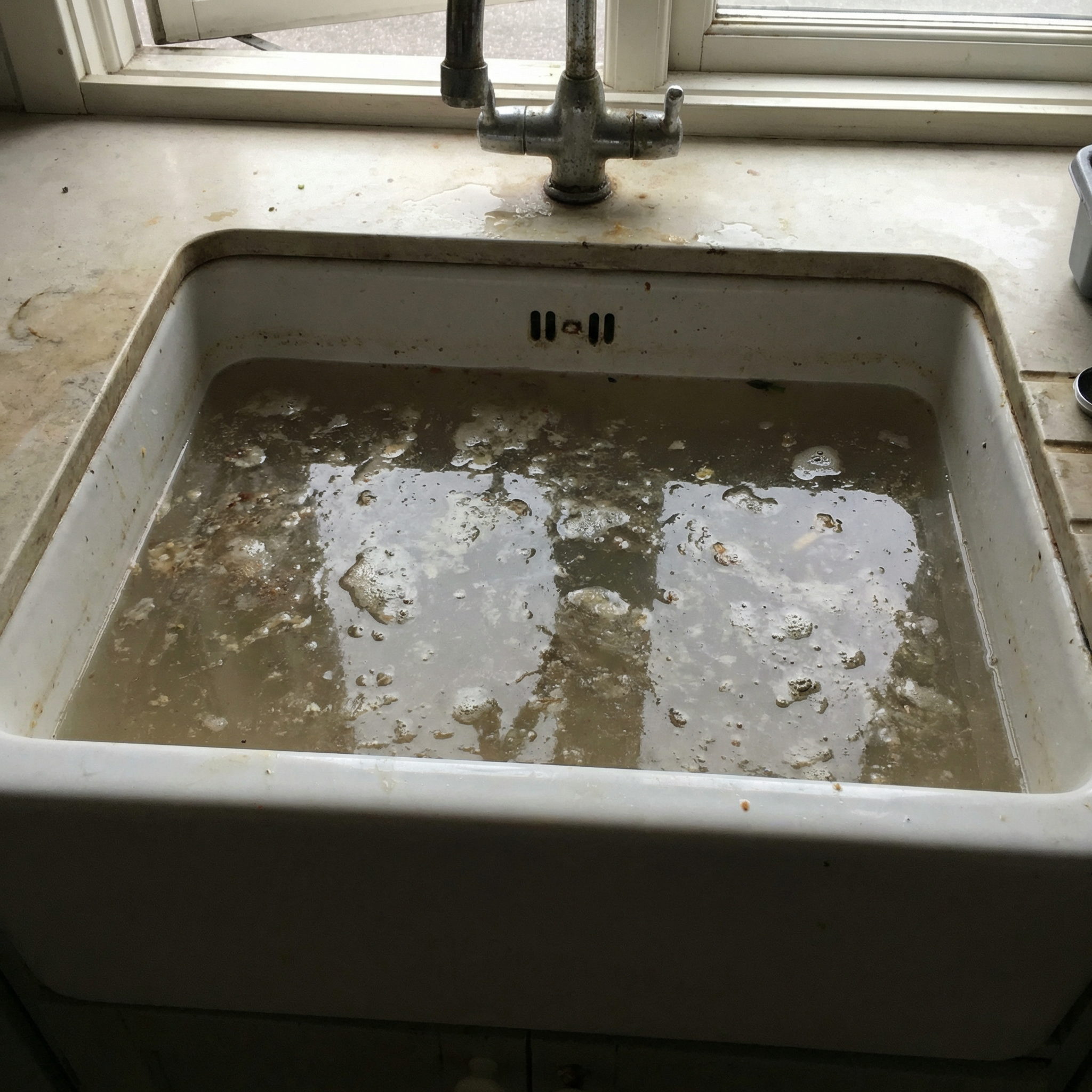 Dirty kitchen sink filled with murky water and debris, with a window above