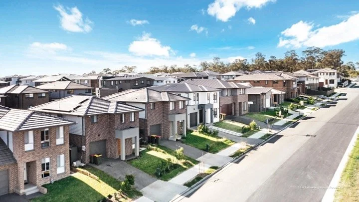 A row of modern suburban houses with brick and concrete exteriors on a sunny day, featuring manicured lawns and driveways in a residential neighborhood.