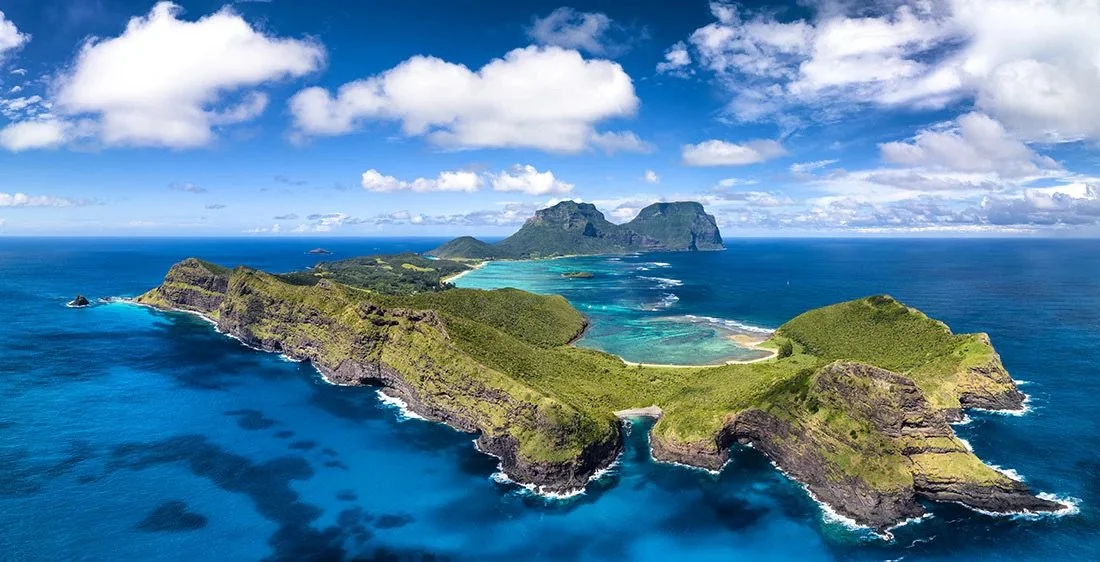 Aerial view of a lush, green island surrounded by turquoise and deep blue ocean waters, under a partly cloudy sky.