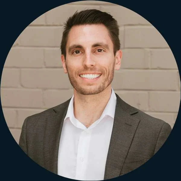 Smiling man in a suit and white shirt against a brick wall backdrop.