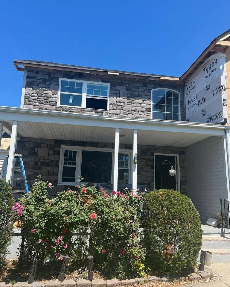 A two-story house under construction with brick and stone exterior, a covered front porch, and a flower bush in front, against a clear blue sky.