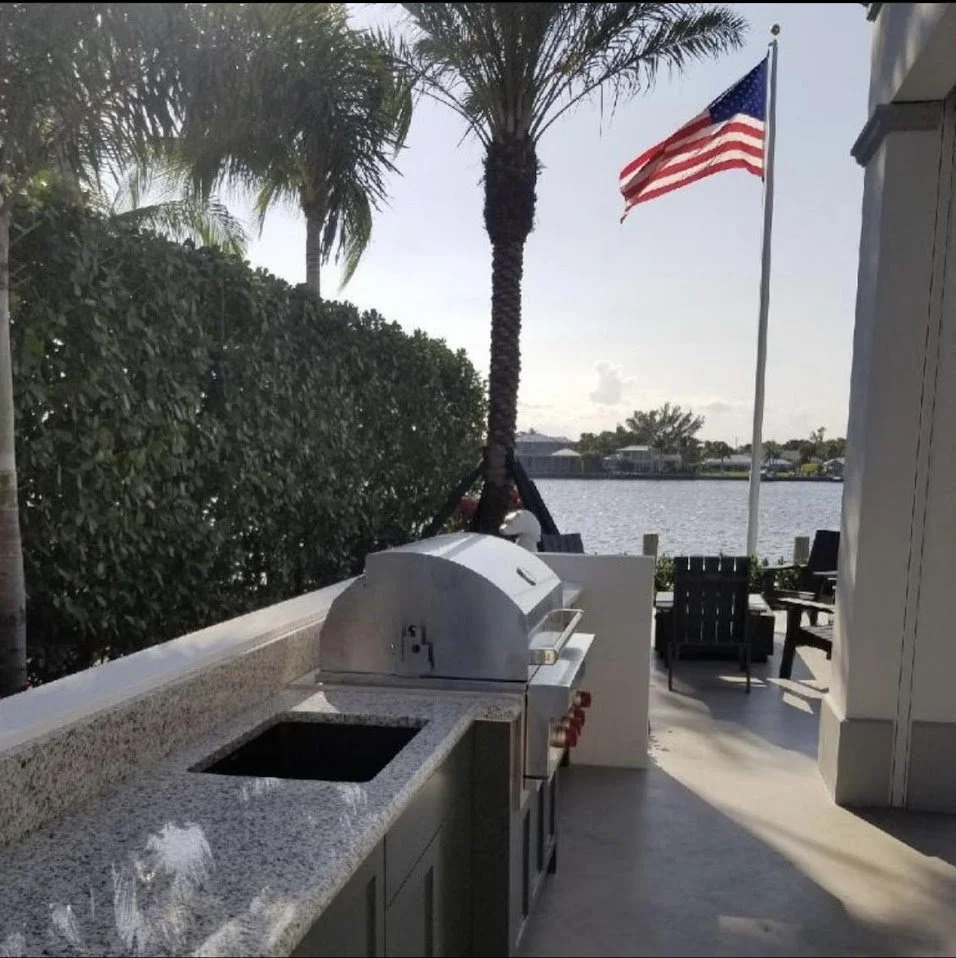 Outdoor view of a patio area with a built-in grill, granite countertop, and seating facing a body of water, with an American flag and palm trees in the background.
