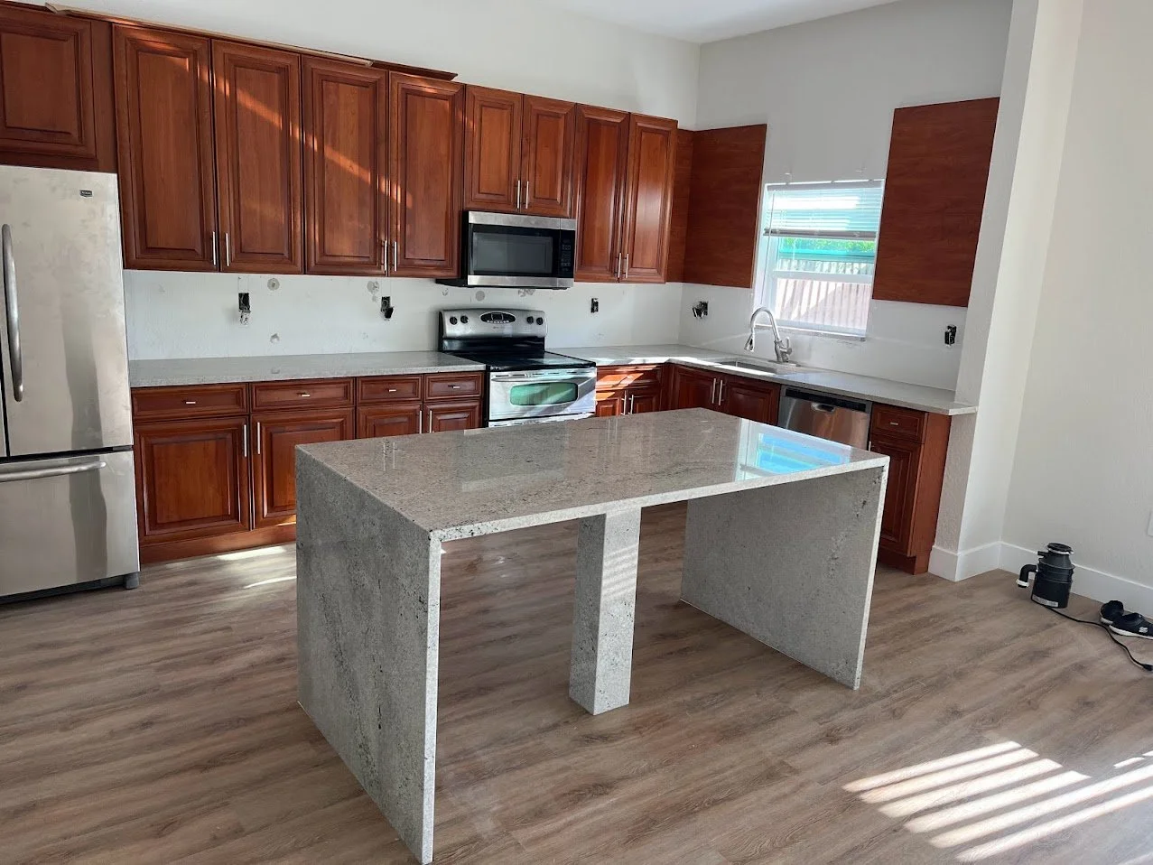 Unfurnished kitchen with wooden cabinets, a stainless steel refrigerator, microwave, stove, dishwasher, a gray granite island, and a window with blinds.