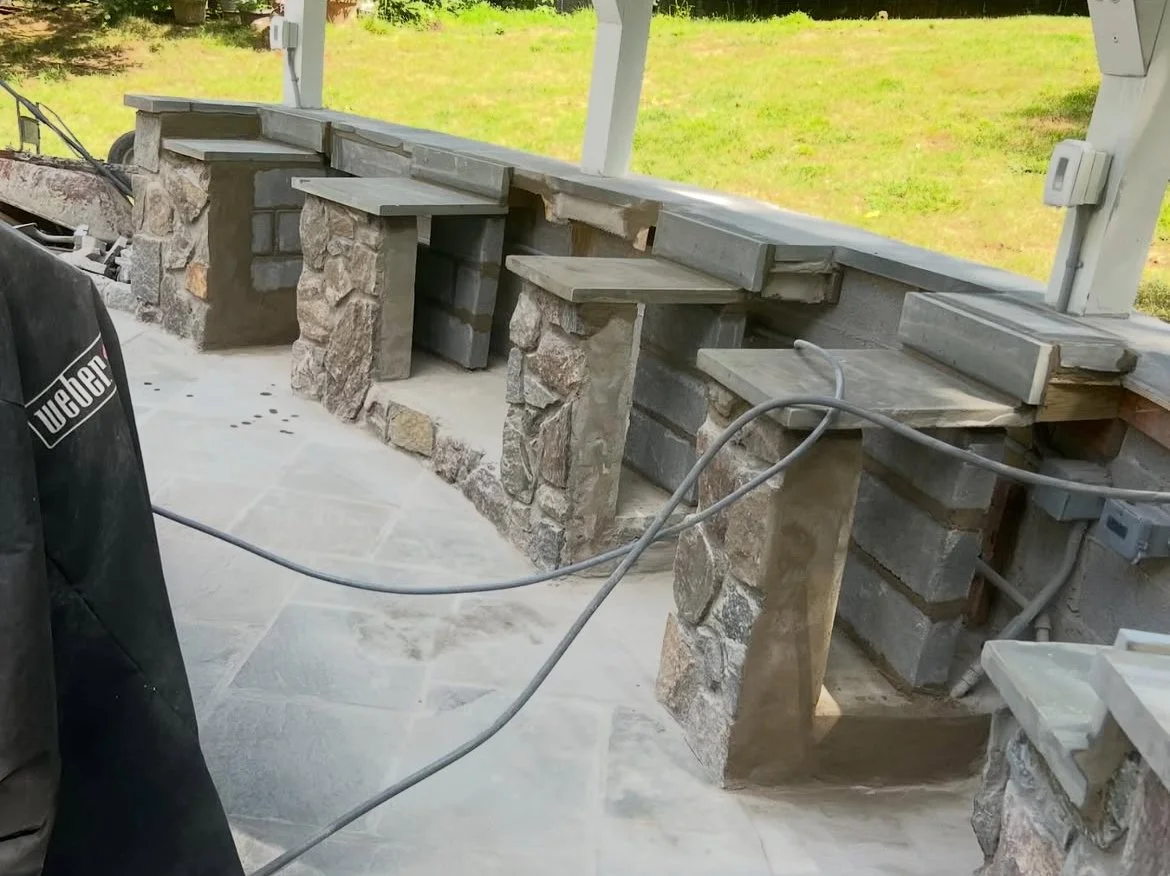Construction of stone and brick outdoor countertop with multiple stone supports and a concrete top, located on a screened porch with electrical outlets and wiring visible.