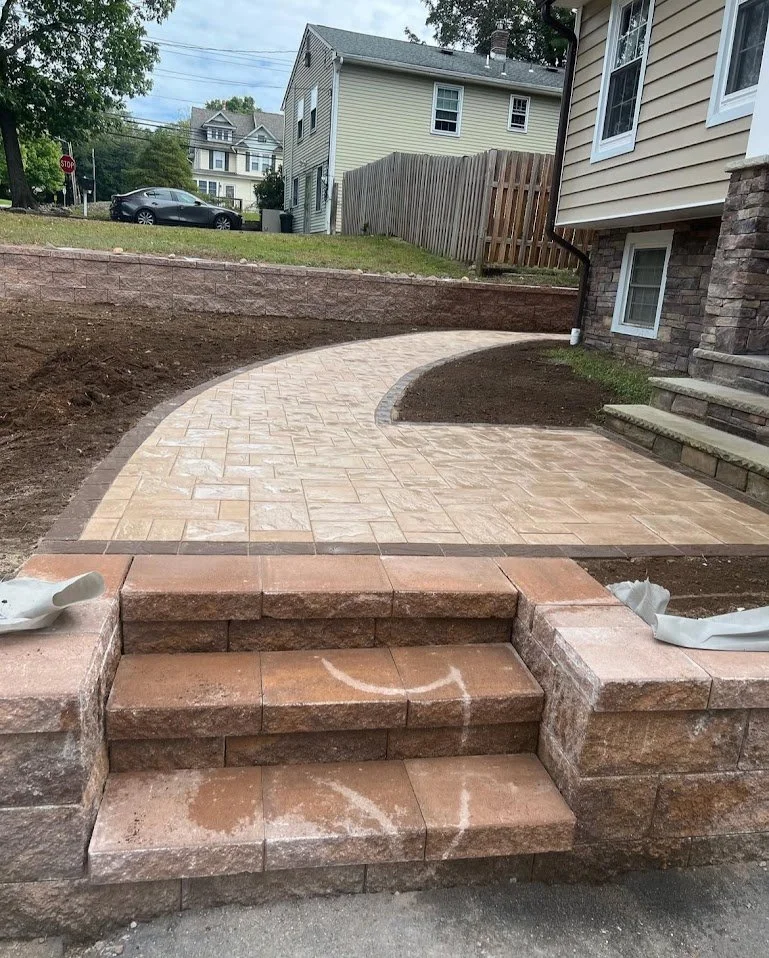 Newly constructed curved stone patio and steps next to a house with beige siding and stone foundation, with soil prepared for additional landscaping.