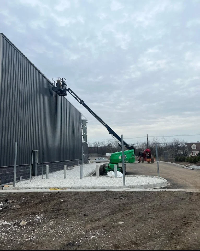 A construction site with a green boom lift worker platform extending towards the top of a large black metal building, surrounded by gravel and unfinished ground.