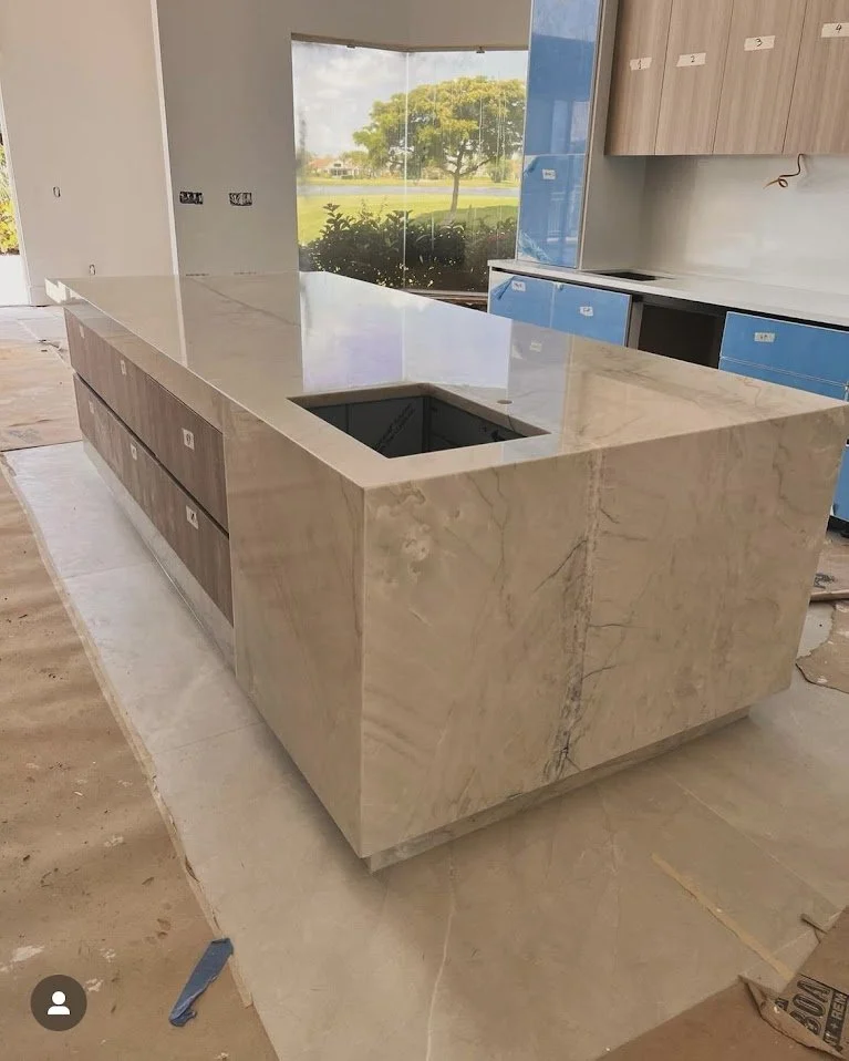 A modern kitchen island with a beige marble countertop and base, featuring an empty cutout for a cooktop, in a room with large windows overlooking a green outdoor scene and incomplete cabinetry.