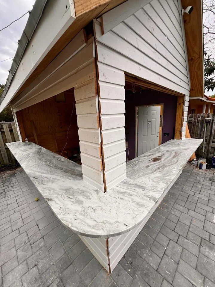 Exterior of a small building under construction, featuring a marble countertop with a gray and white pattern extending around the corner, and unfinished siding with white paint and exposed wood framing.