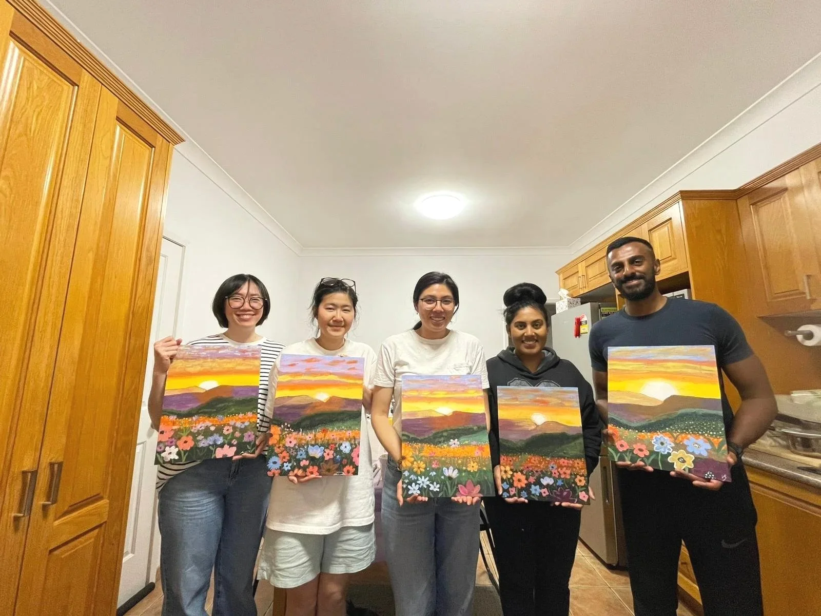 Group of five people in a kitchen holding colorful landscape paintings of a sunset over mountains and flowers.