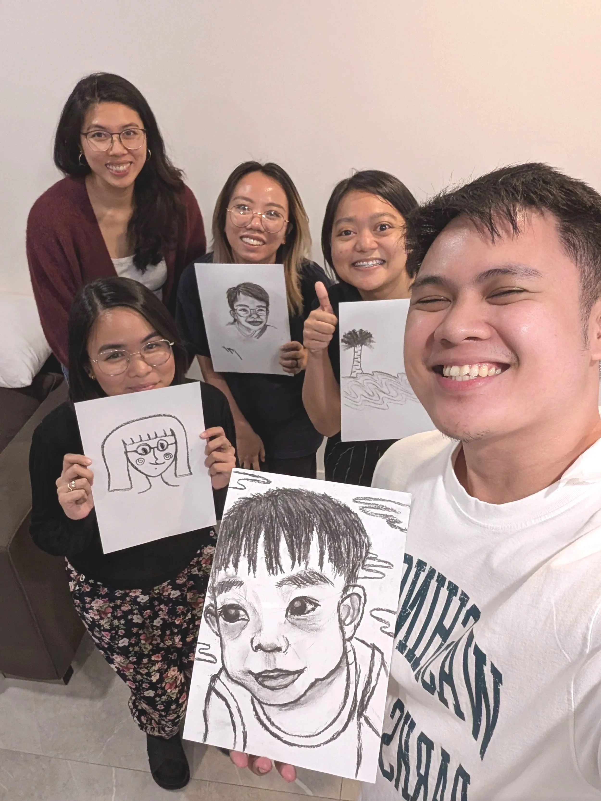Group of five women and one man holding up sketches of portraits, smiling and standing in a room with a white wall.