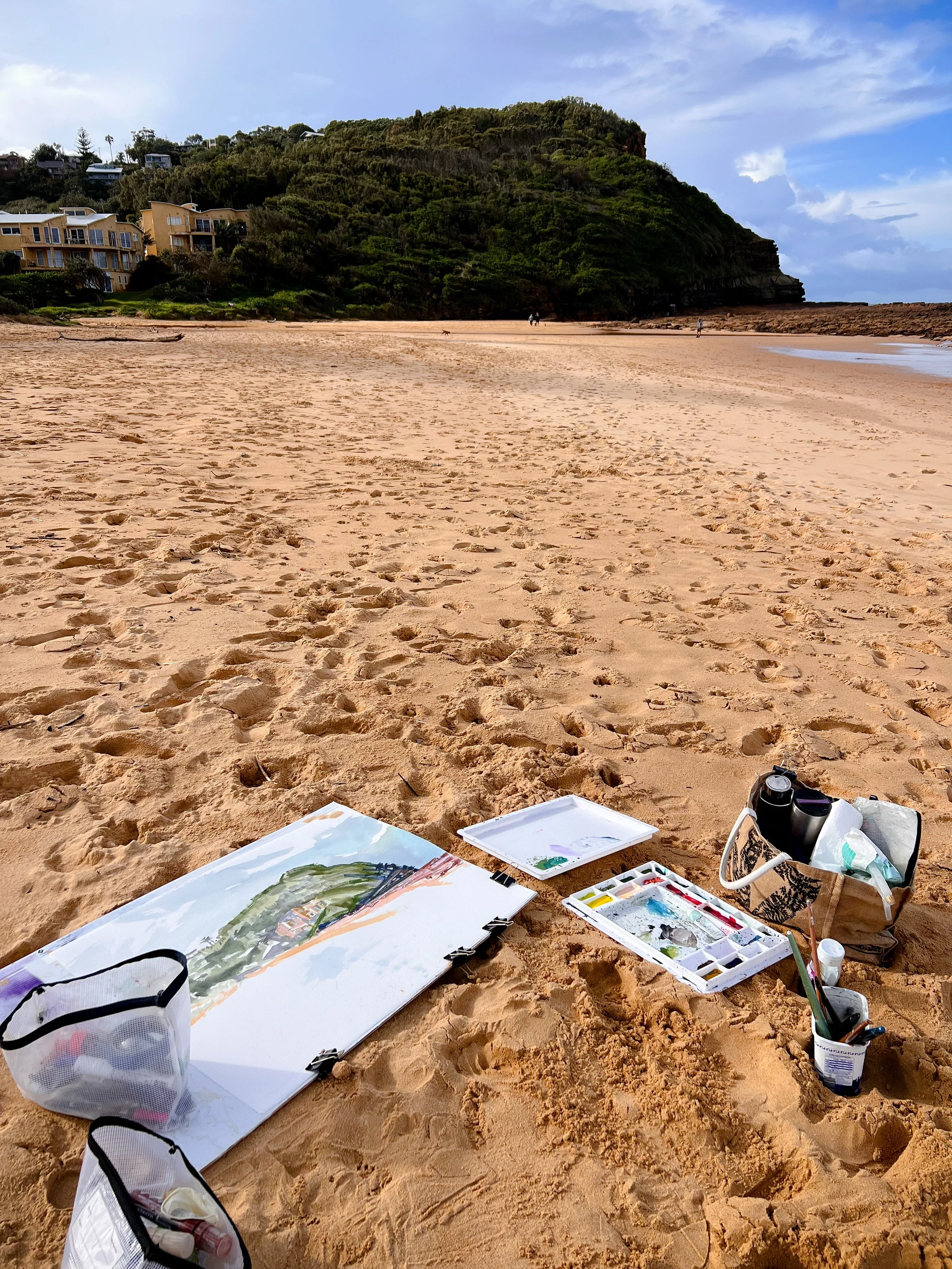 Beach scene with painting supplies on sand, including a full sheet of watercolour paper with a landscape painting, watercolor palette, and bags, with houses and a green hillside in the background.