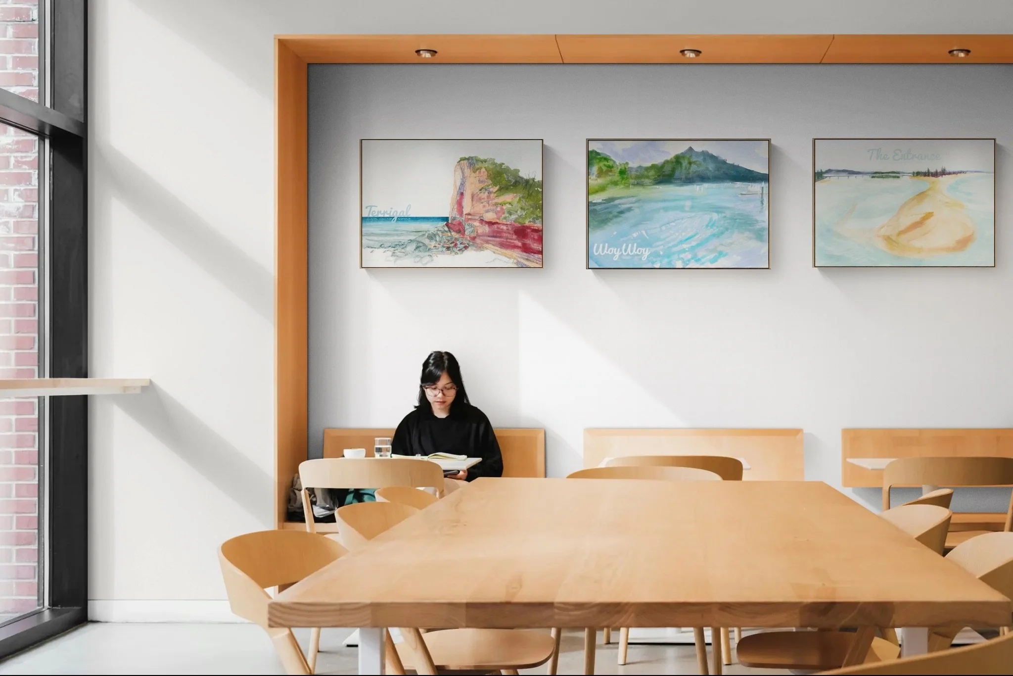 A woman with black hair and glasses sits at a table in a modern cafe, reading a book with a glass of water and a small bowl nearby. The cafe has wooden tables and chairs, with three framed watercolor paintings of landscapes on the wall behind her. Large windows let in natural light.
