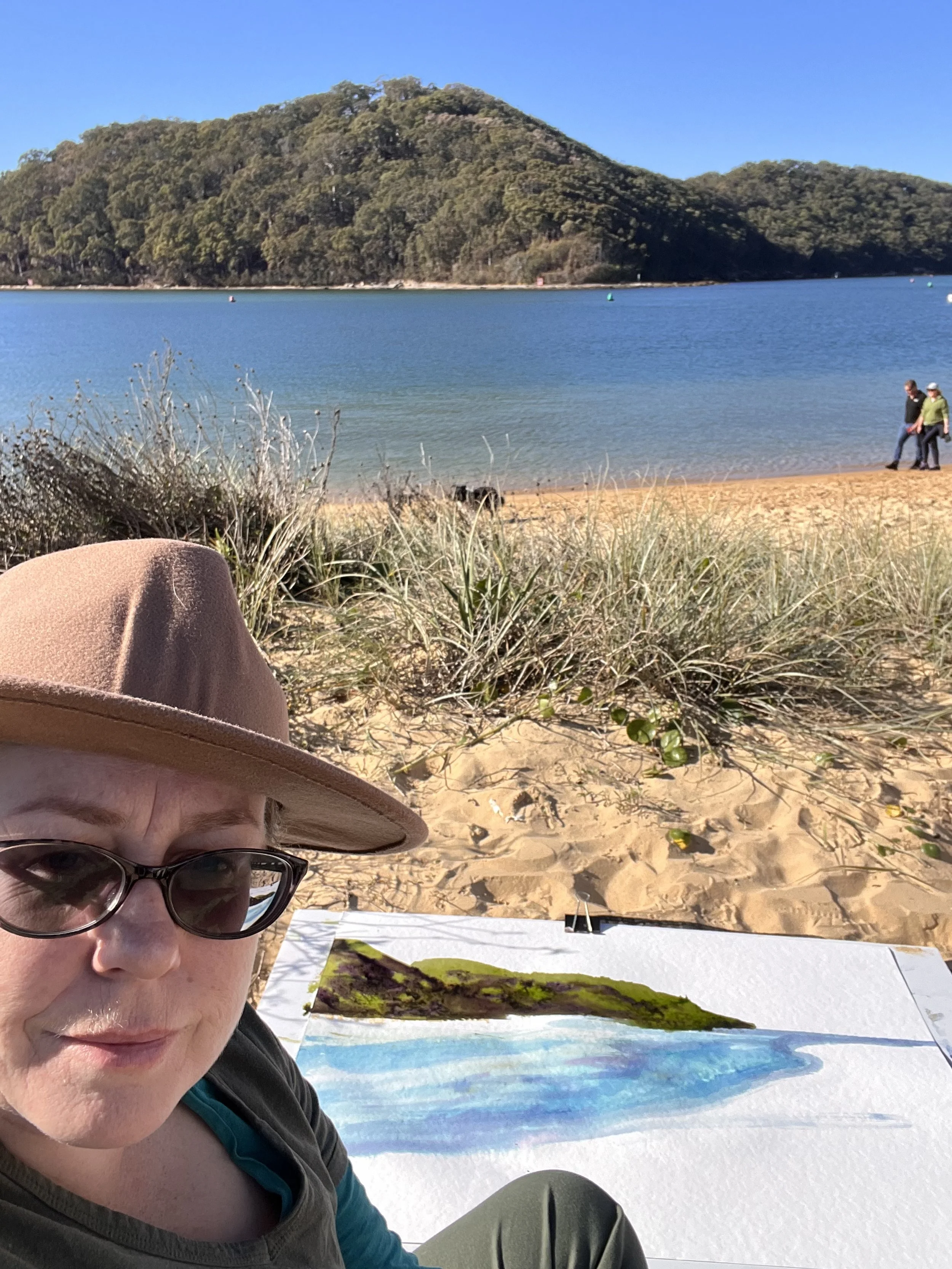 Maz wearing a hat painting a landscape by the sea with a landform in the background and two people walking on the shore.