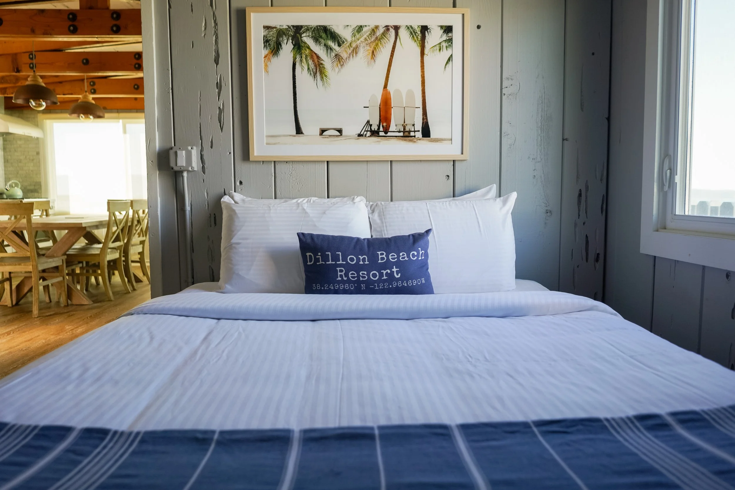 A bright bedroom at Dillon Beach Resort with a white-striped duvet, a navy blue accent pillow, and tropical palm tree wall art. In the background, a warm wooden dining area is visible through an open doorway.
