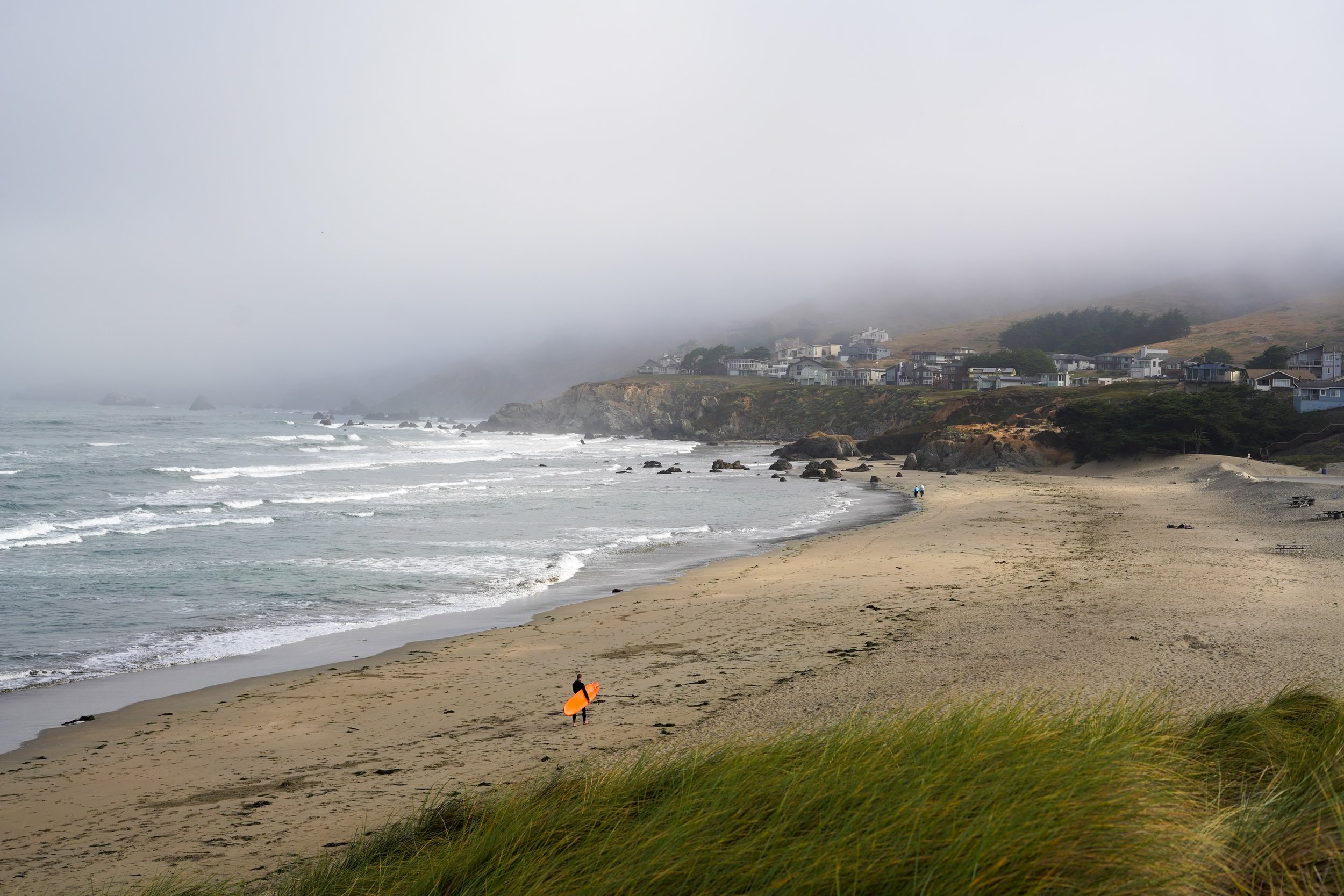 A foggy beach scene with houses on a hillside, a person carrying an orange surfboard walking on the sand, and ocean waves crashing against rocks.