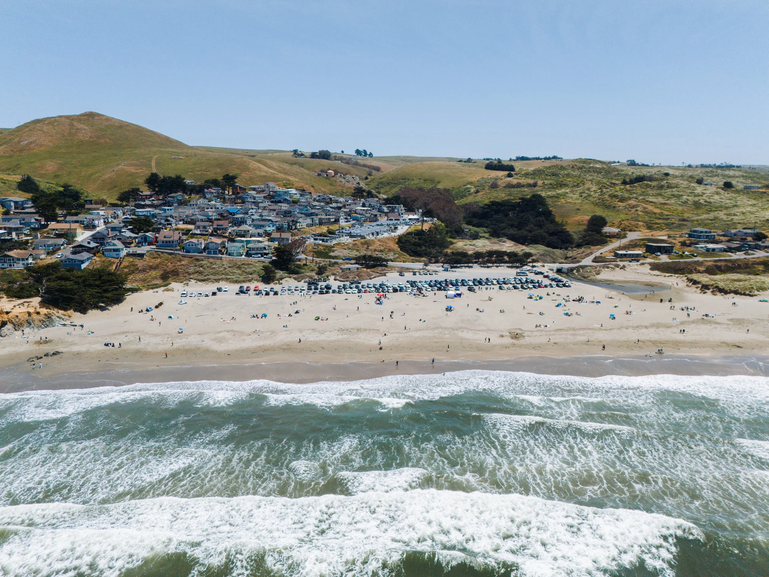 Aerial view of a beach with people, waves, and parked cars, with hilly terrain and houses in the background under a clear blue sky.