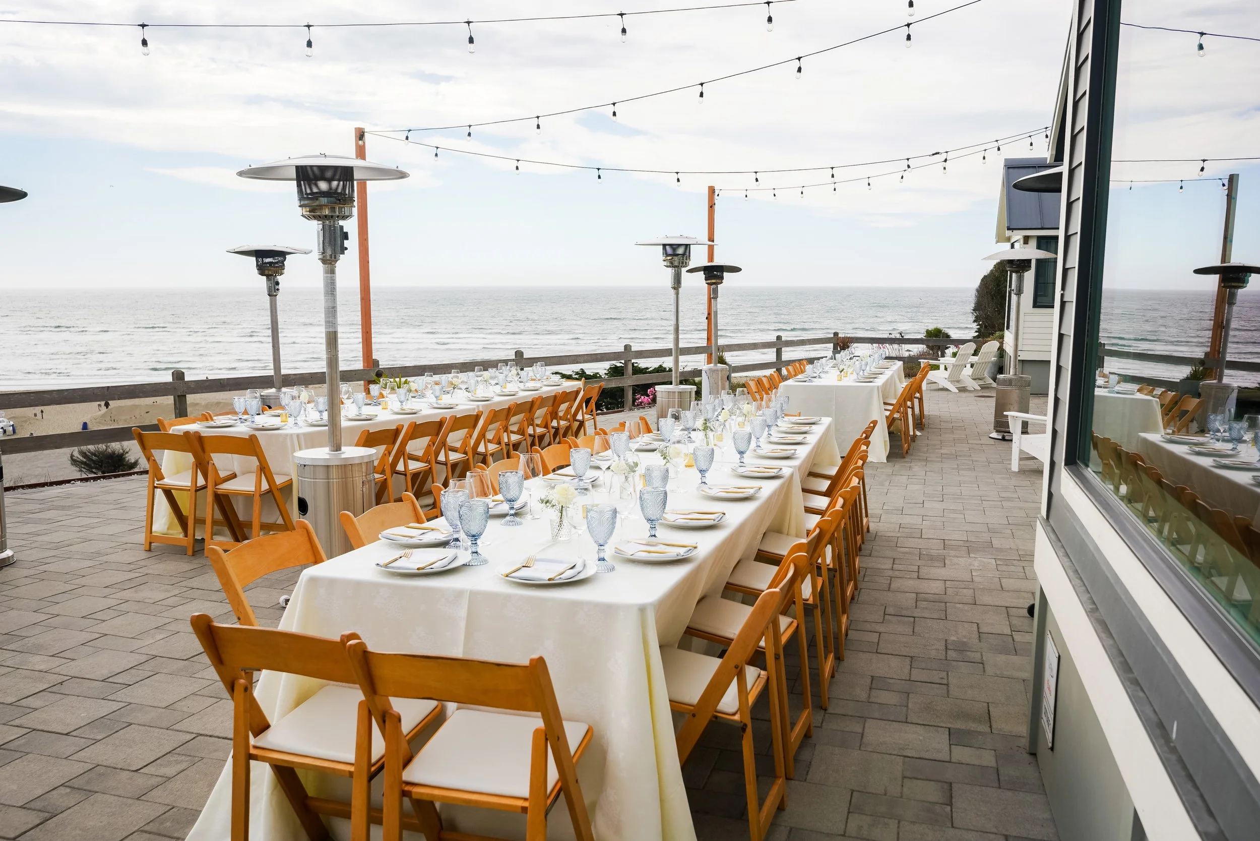 Outdoor dining setup on a patio with long tables covered with white tablecloths, set with glassware, plates, and utensils, overlooking the beach and ocean with string lights overhead.