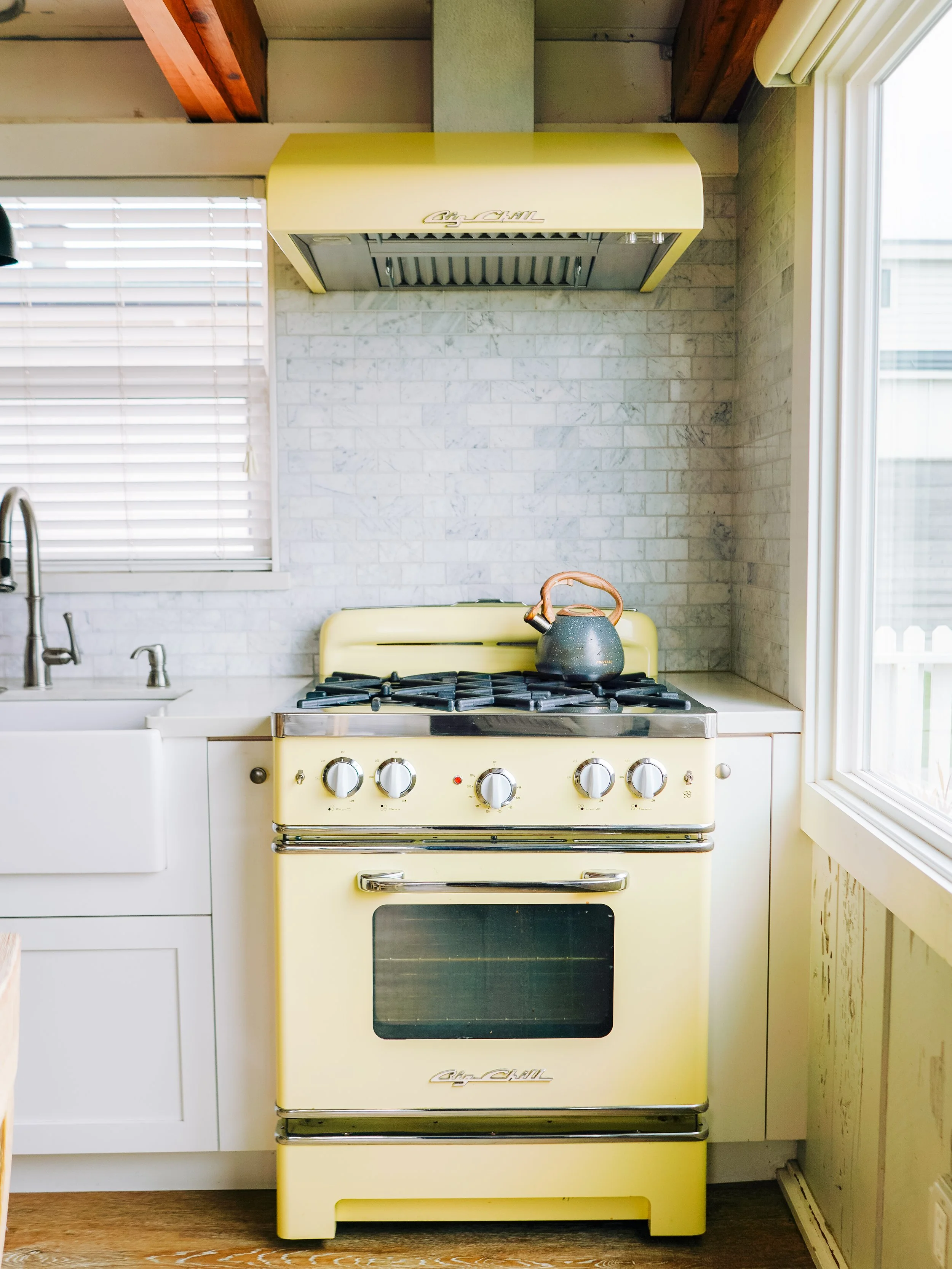 Detail of a pale yellow retro-style Big Chill gas range and matching vent hood against a white marble tile backsplash in a coastal kitchen.