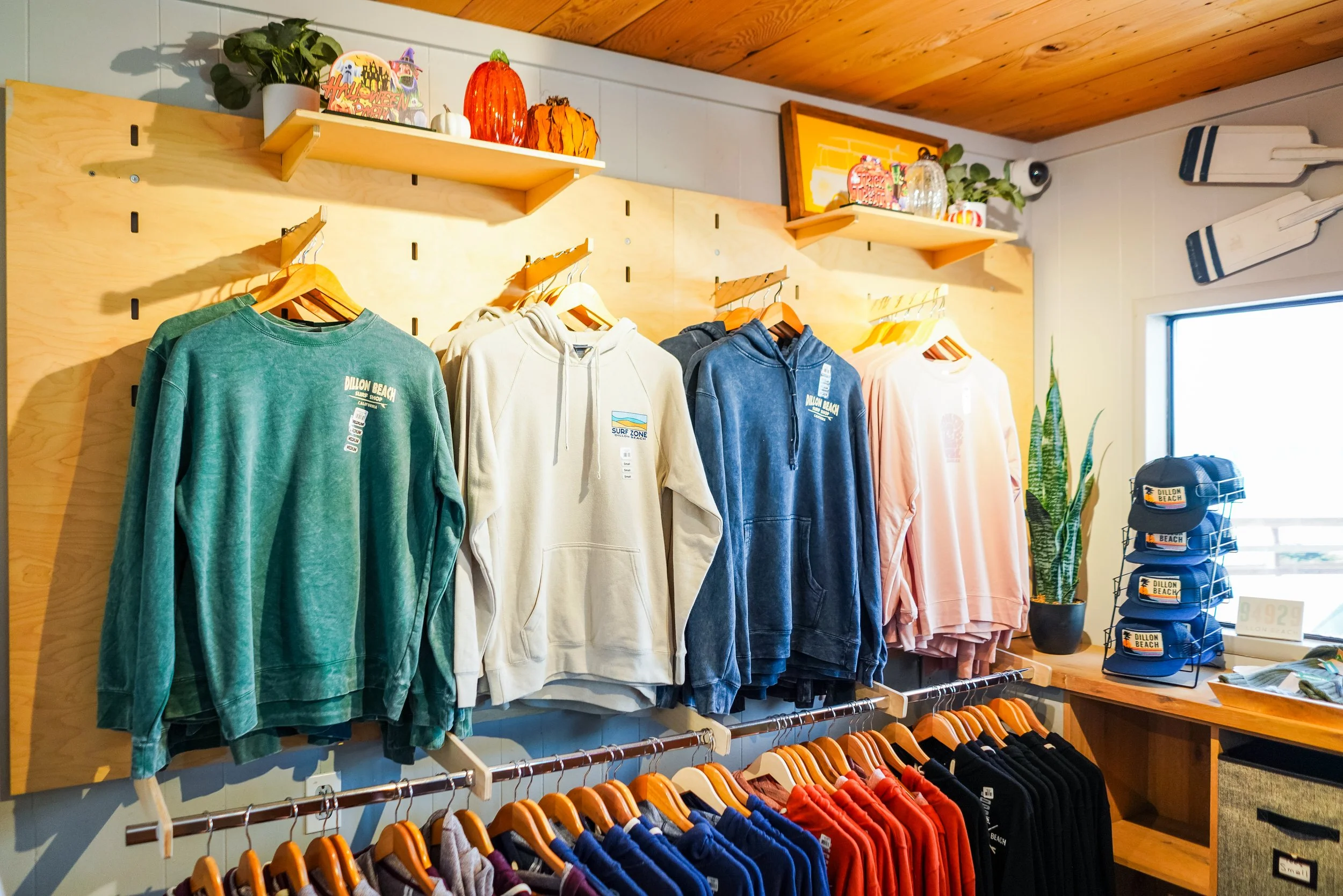 Clothing store display with hoodies on hangers in green, cream, blue, and pink, and T-shirts below in various colors. Shelves above hold decorative pumpkins, plants, and small Halloween signs, with a wooden ceiling and large window in the background.