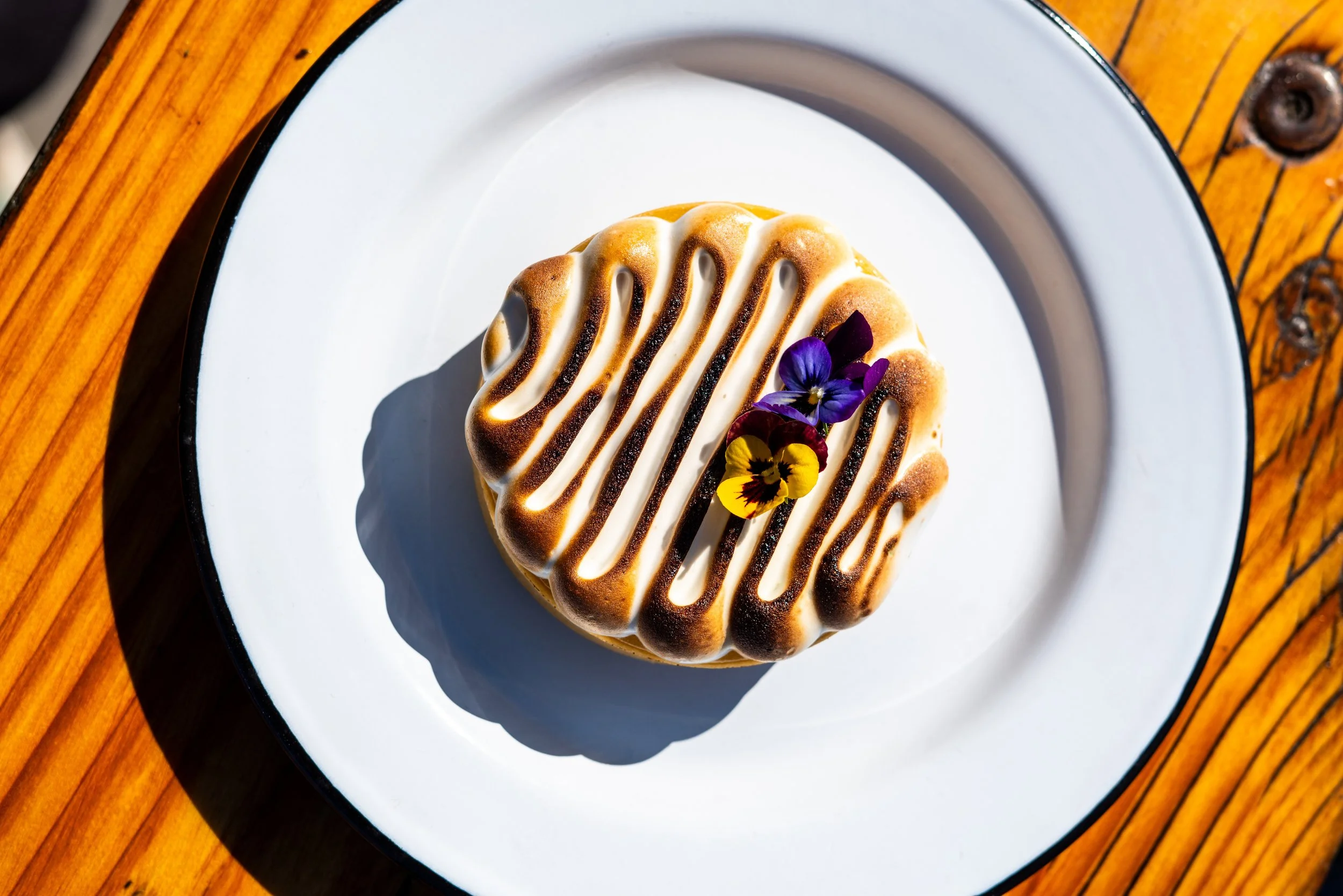 Dessert with white and dark drizzle, garnished with edible flowers on a white plate with a black rim, placed on a wooden table.