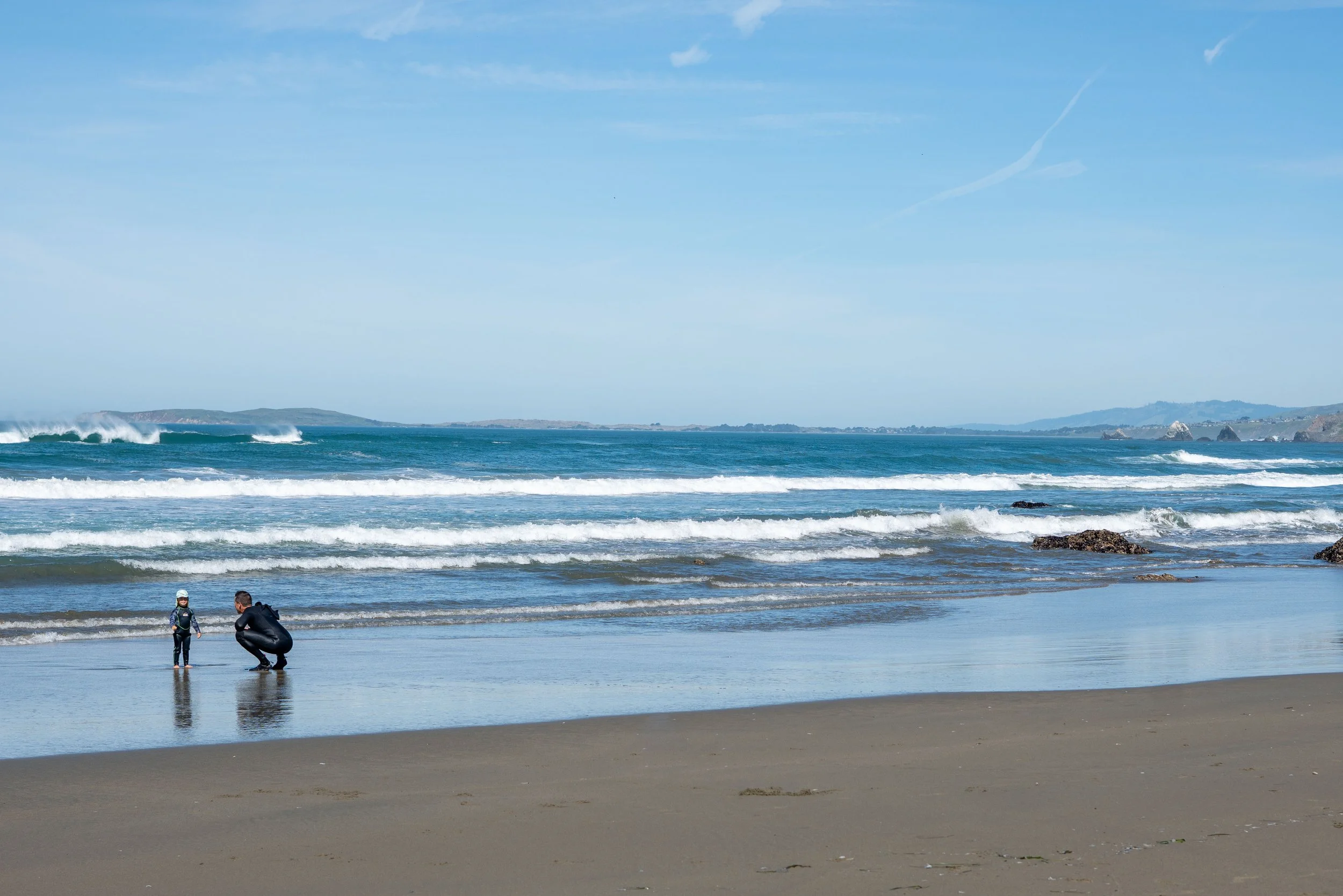 A beach with people in wetsuits preparing for surfing, ocean waves, and a distant shoreline with hills under a partly cloudy sky.