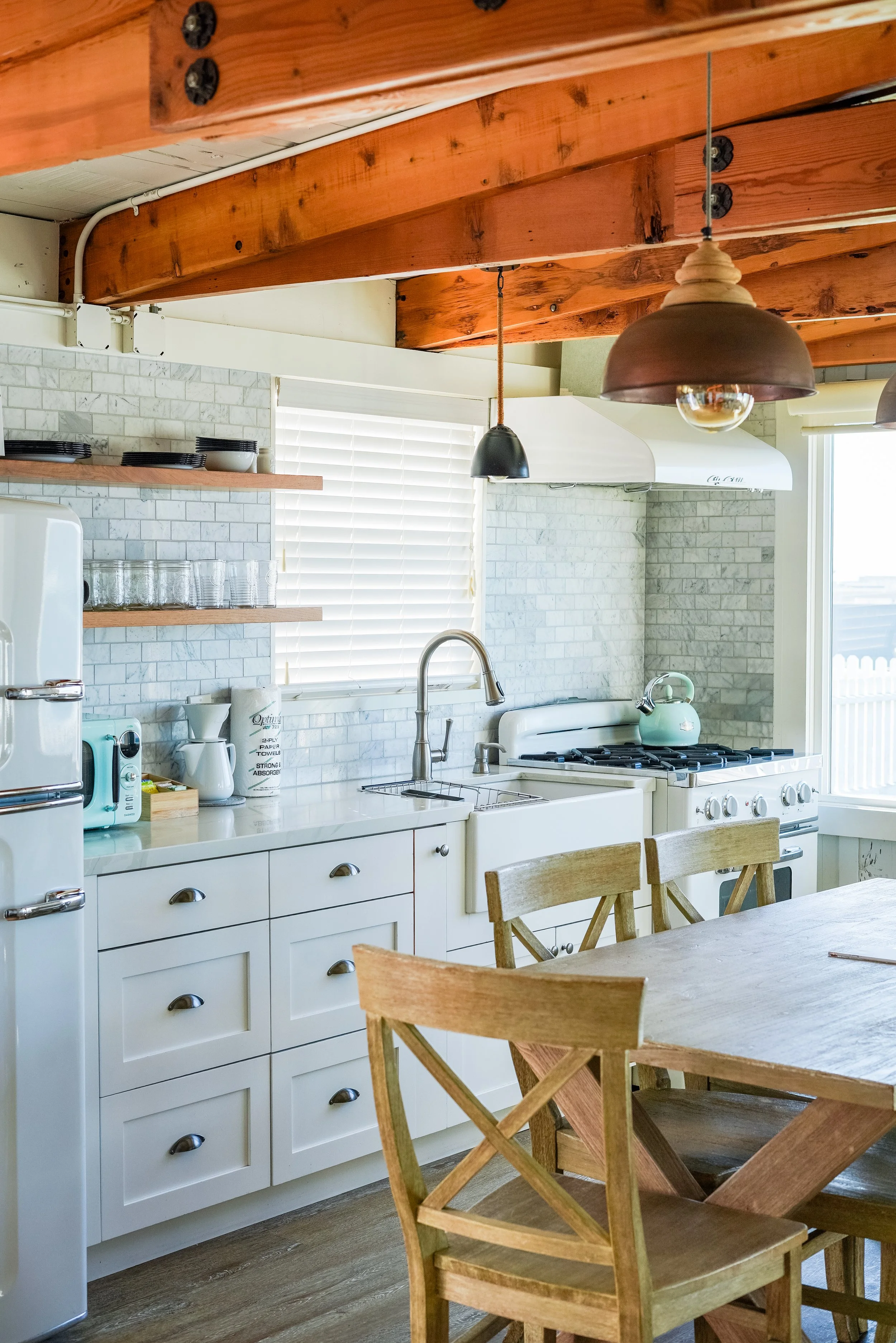 A bright kitchen with white shaker cabinets, a large farmhouse sink, and white marble tile backsplash. The space features exposed wood ceiling beams, a vintage-style white gas range, and a wooden dining table with cross-back chairs.