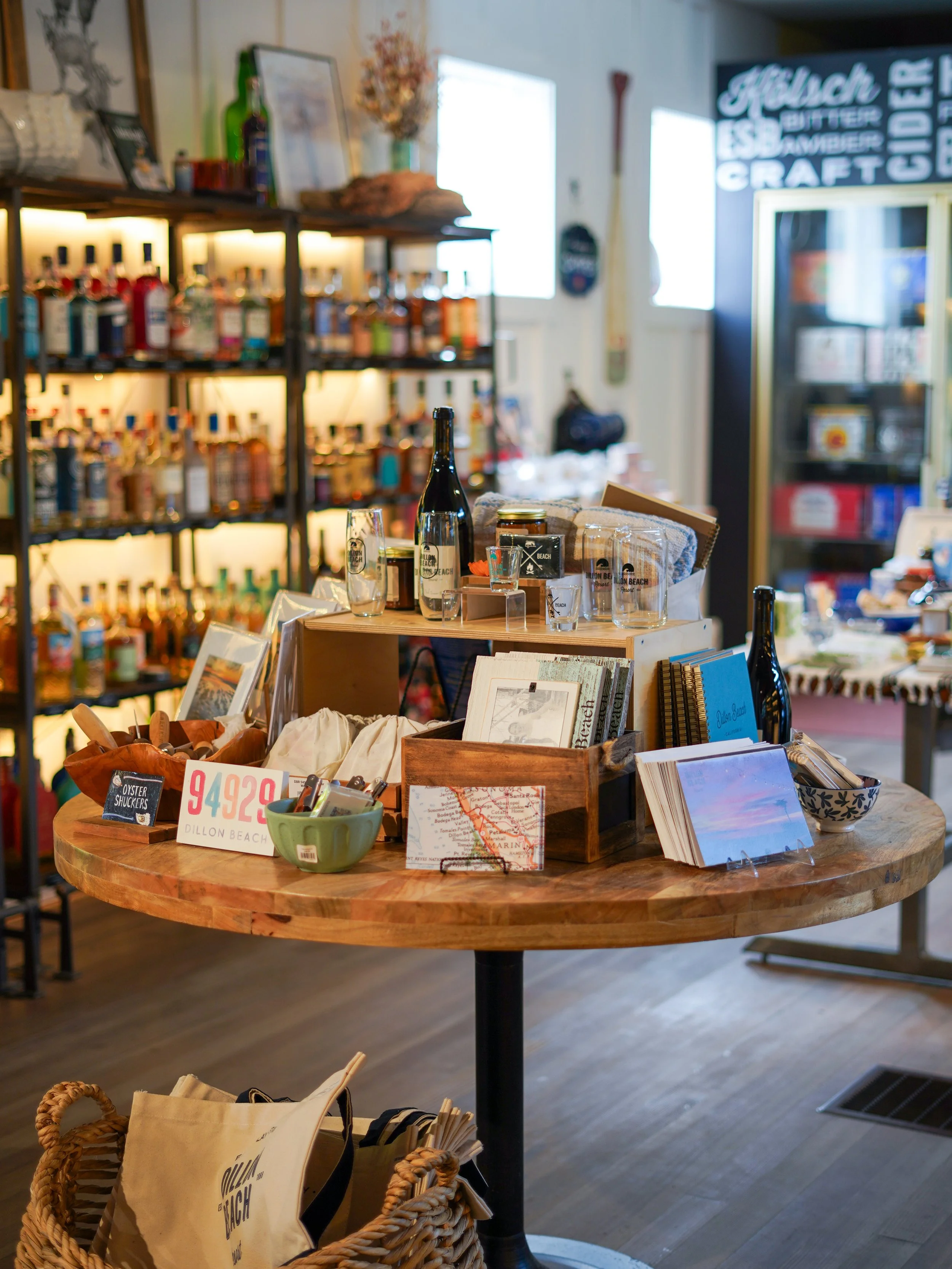 Circular wooden table displaying various pamphlets, postcards, and decorative items, with a basket containing tote bags in the foreground and shelves filled with bottles in the background, inside a cozy shop or cafe.